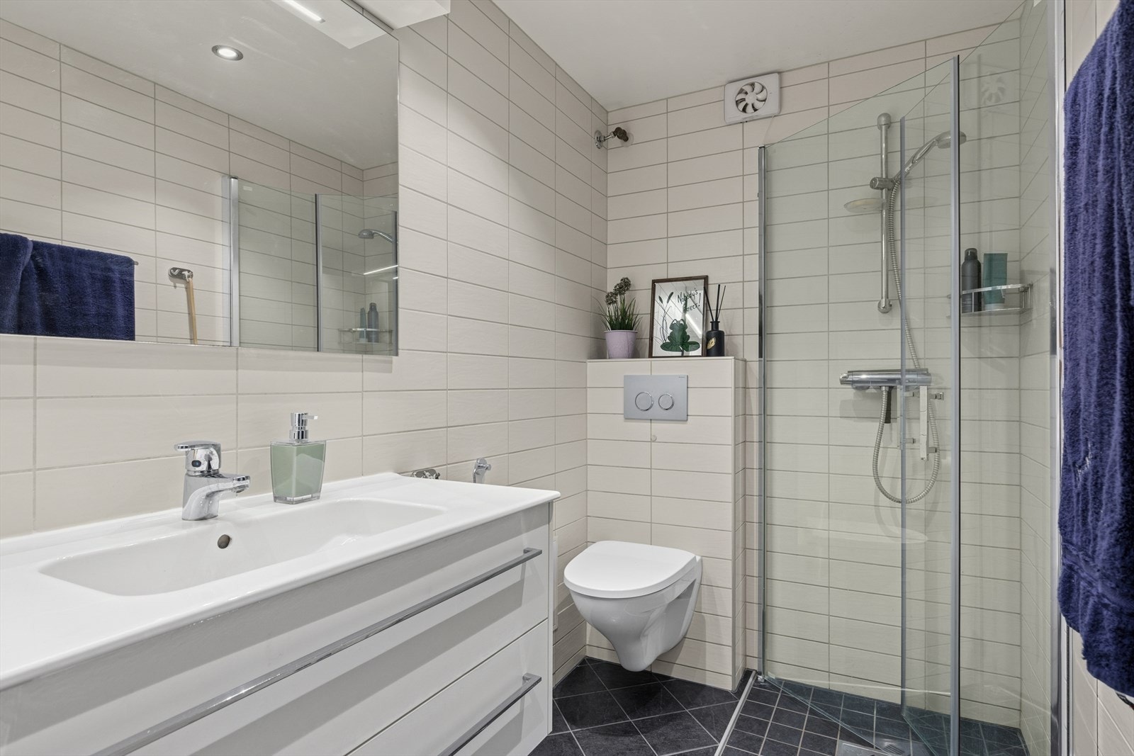 Flislagt bad/wc med servantskap, speil, vegghengt toalett, svingbare dusjdører og opplegg til vaskemaskin/tørketrommel. Galleribilde