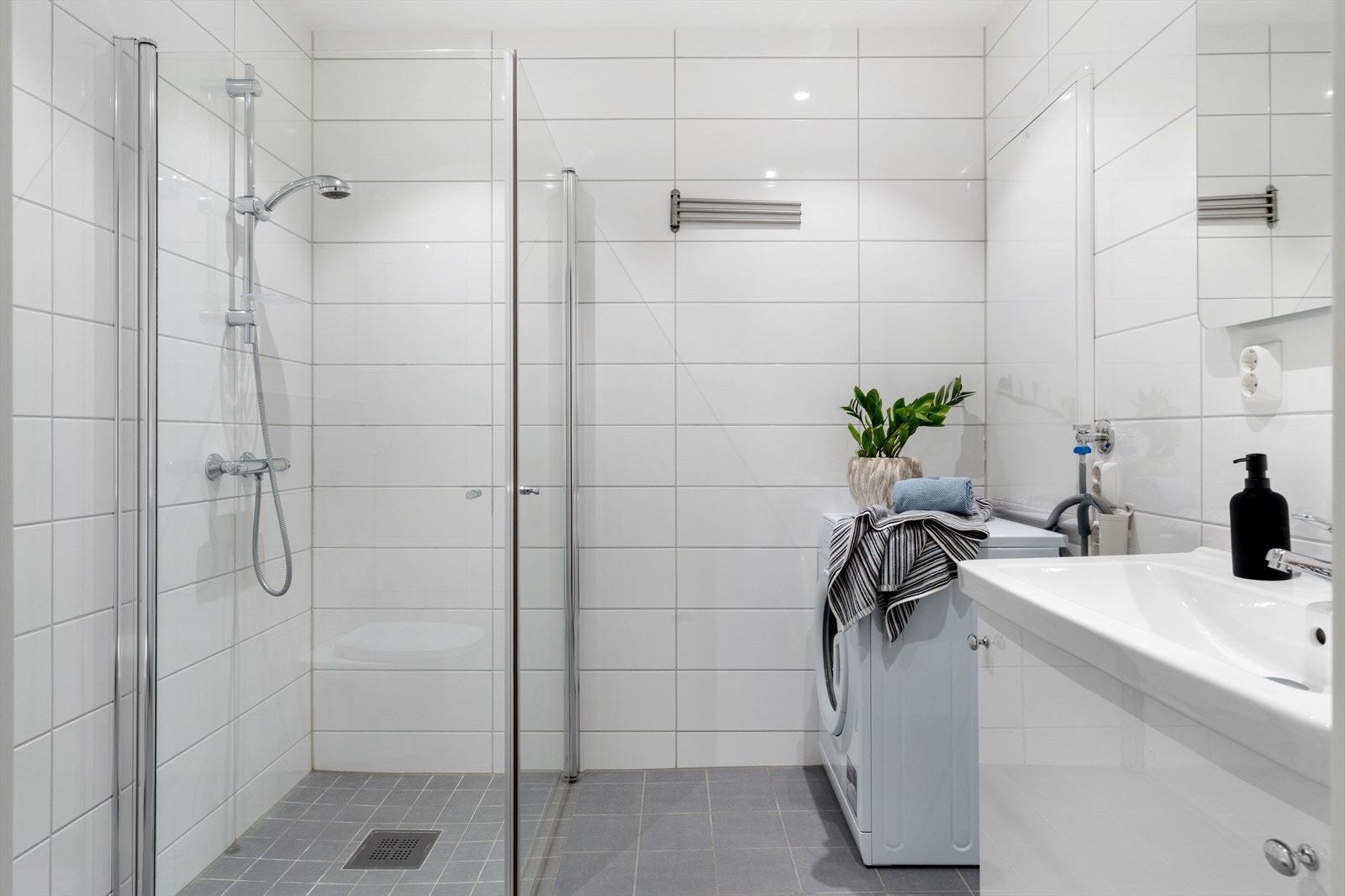 Badet er innredet med servant i møbel, speil med lys, vegghengt toalett, dusj med glassdører og opplegg for vaskemaskin. Galleribilde