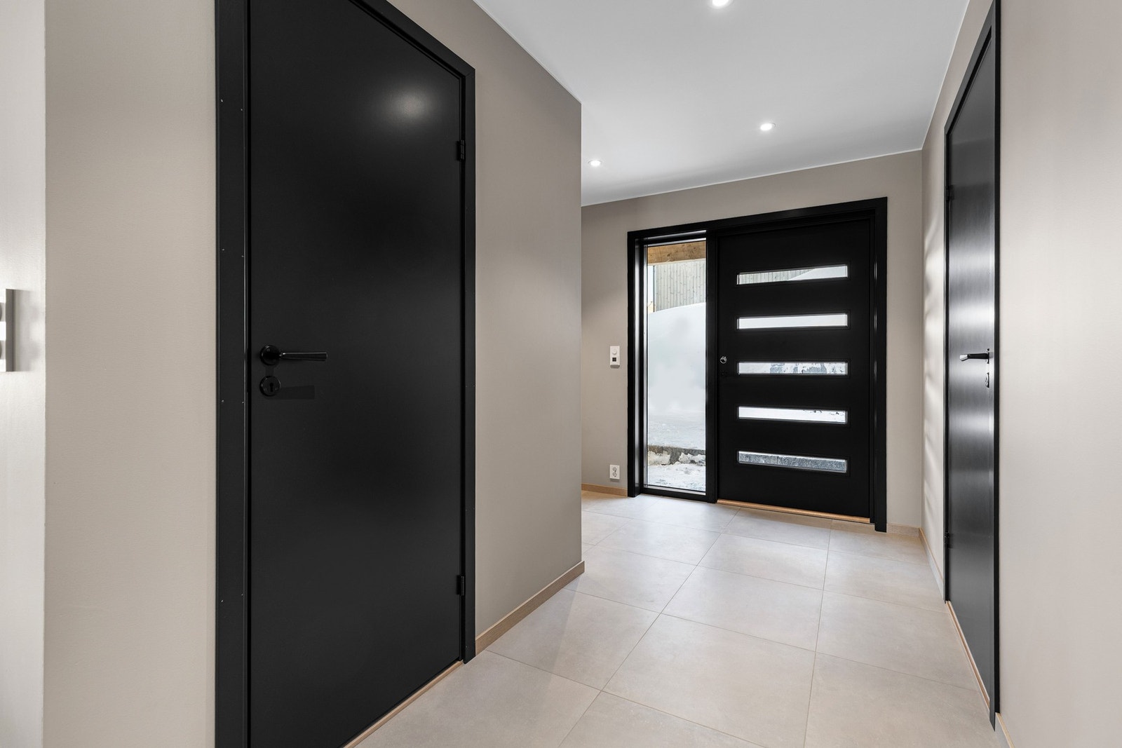 Den sorte ytterdøren med fem horisontale glassfelt ønsker deg velkommen inn i en innbydende entre med 60x60 cm fliser og varmekabler i gulvet. Galleribilde