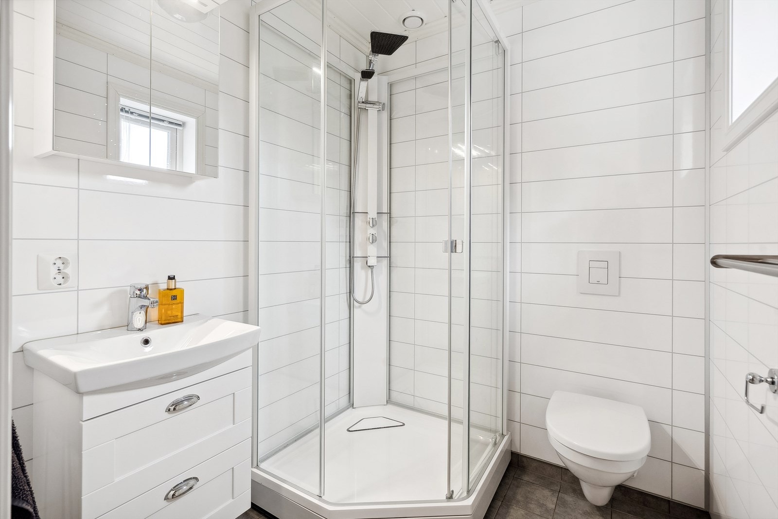 Badet i 1.etasje er av mindre størrelse. Badet inneholder dusjkabinett, servant med skuffer, overskap med speil og vegghengt toalett. Galleribilde