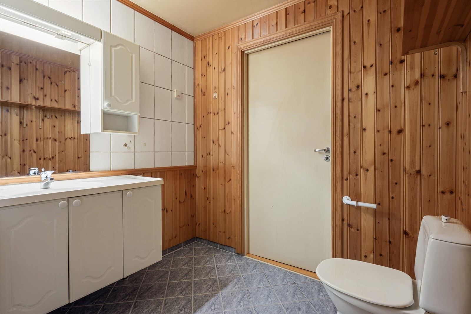 Vegger med fliser og trepanel, samt gulv med fliser. Badet er innredet med klosett, servant med innredning og badekar med dusjutstyr og forheng Galleribilde