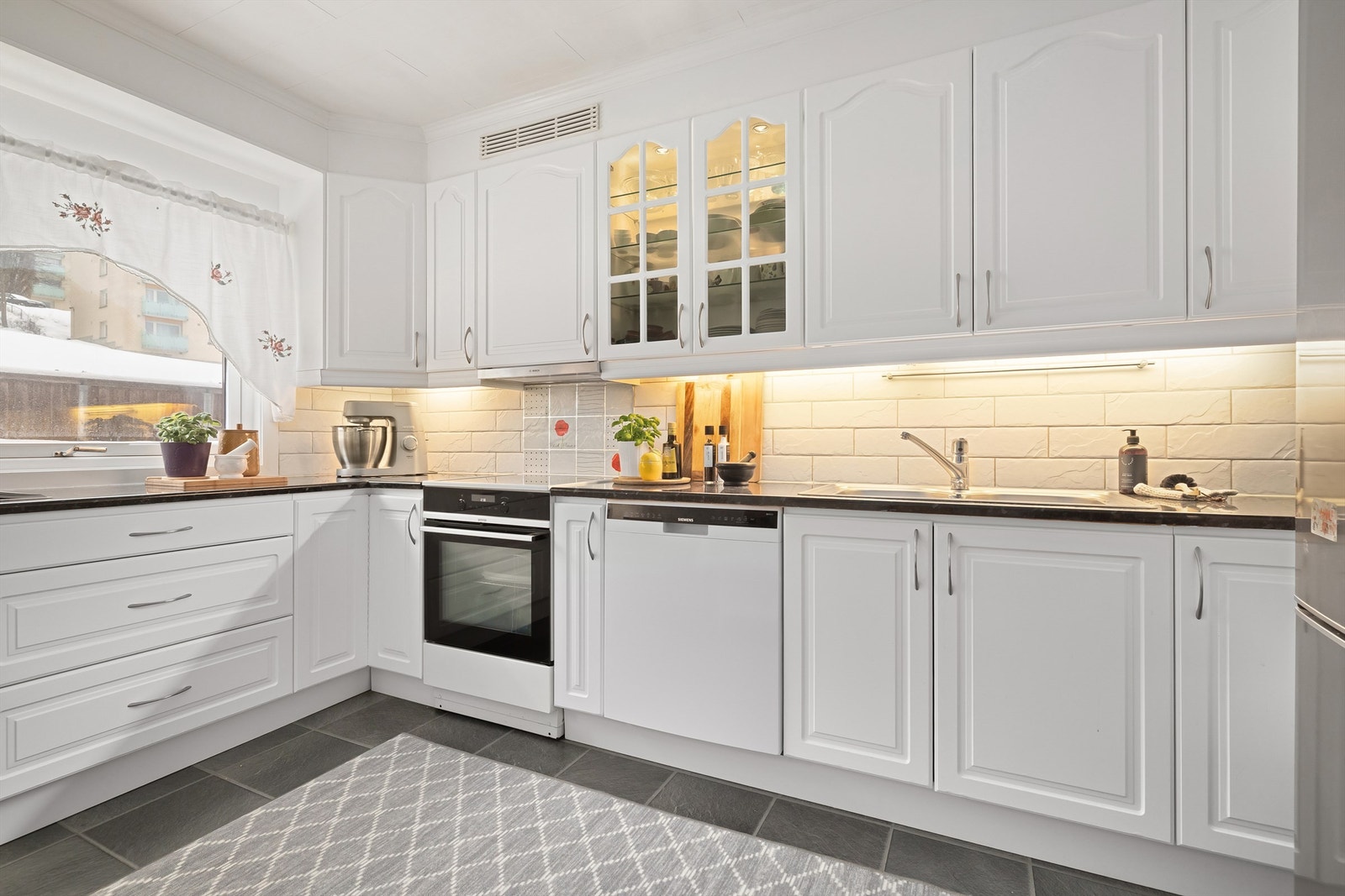 Kjøkkeninnredningen består av profilerte fronter med fliser mellom laminert benkeplate og overskap, nedfelt vask, samt benkeskapsbelysning og stikkontakter under overskap. Galleribilde