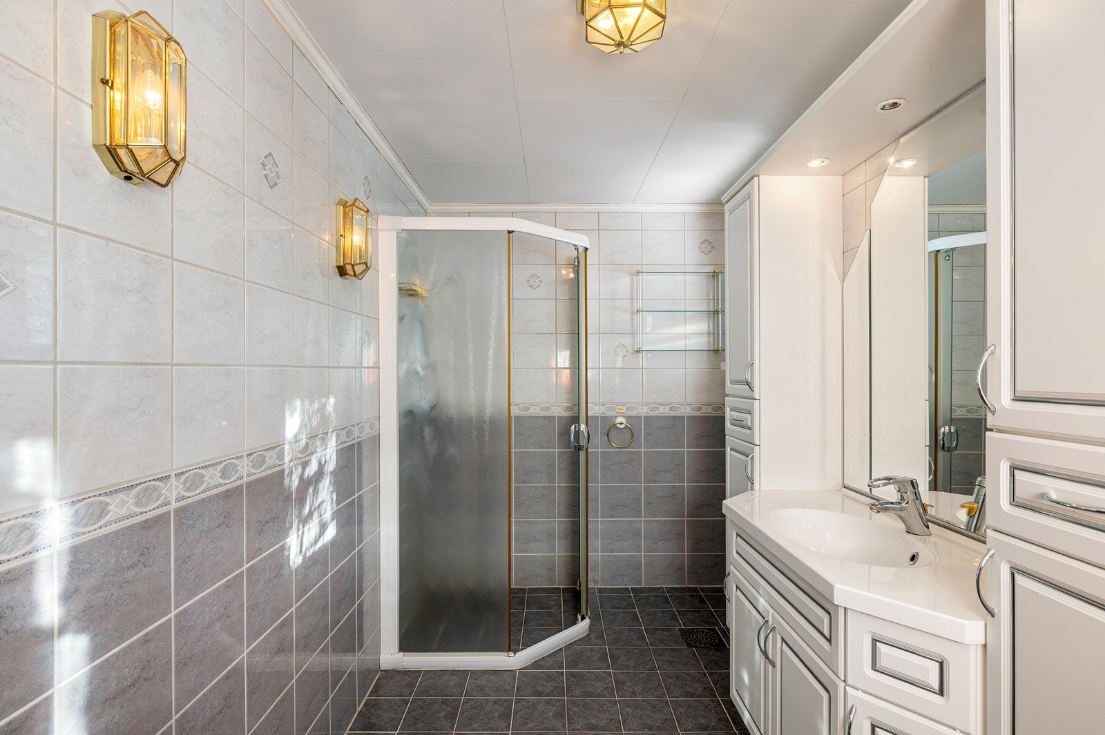 Fra gangen er det inngang til et flislagt bad. Innredningen består av et underskap/overskap, servant, speil, lys og en dusjsone med dusjing direkte på gulv og vegg. Galleribilde