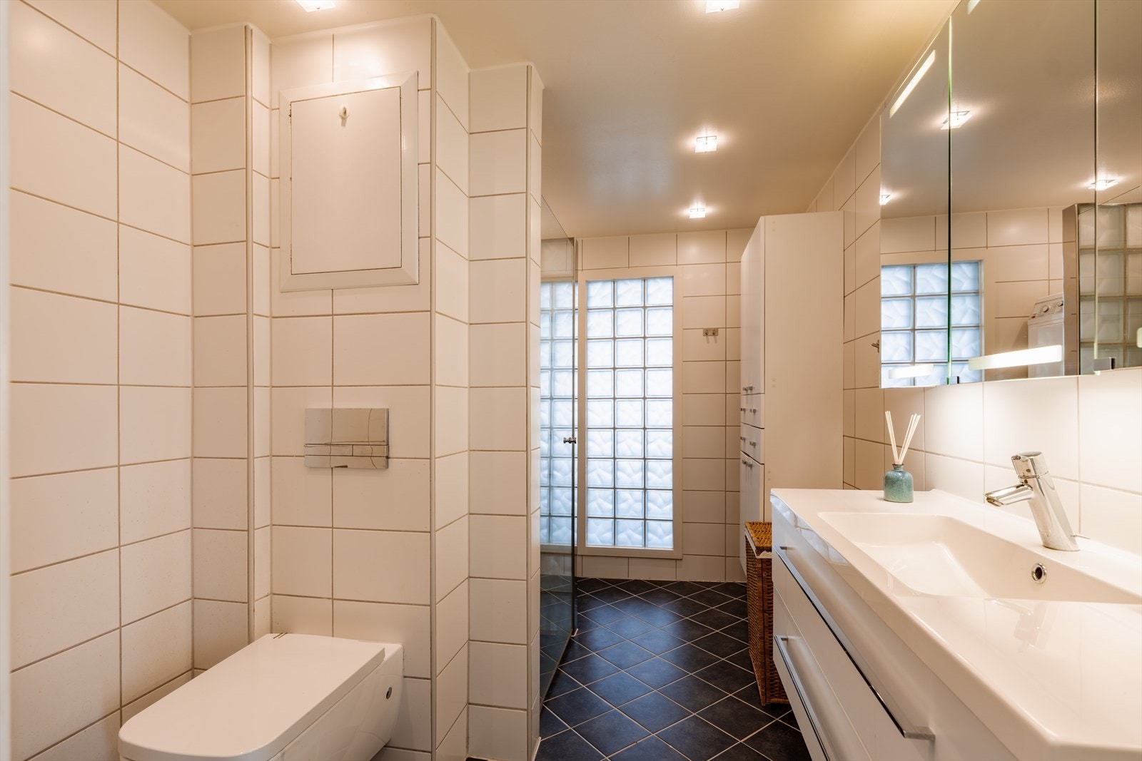 Flislagt bad med varmekabler i gulv. Innredningen består av benkeskap og veggskap med glatte fronter, servant og vegghengt toalett med innebygget sisterne. Galleribilde