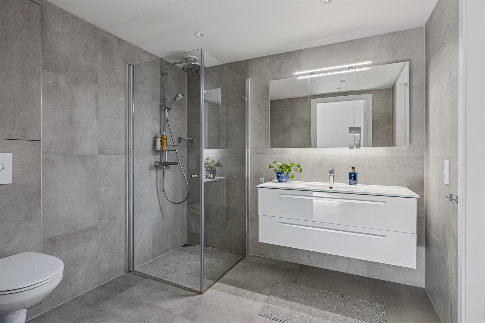 Delikat baderom med vegghengt toalett, dusjhjørne med dører og en servantinnredning med skuffer. Galleribilde