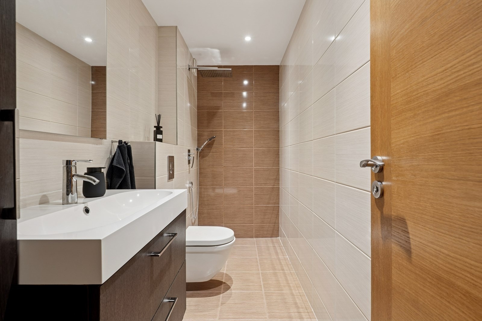 Moderne og stilrent baderom med vegghengt toalett, romslig dusjsone og delikate fliser i varme toner. Galleribilde