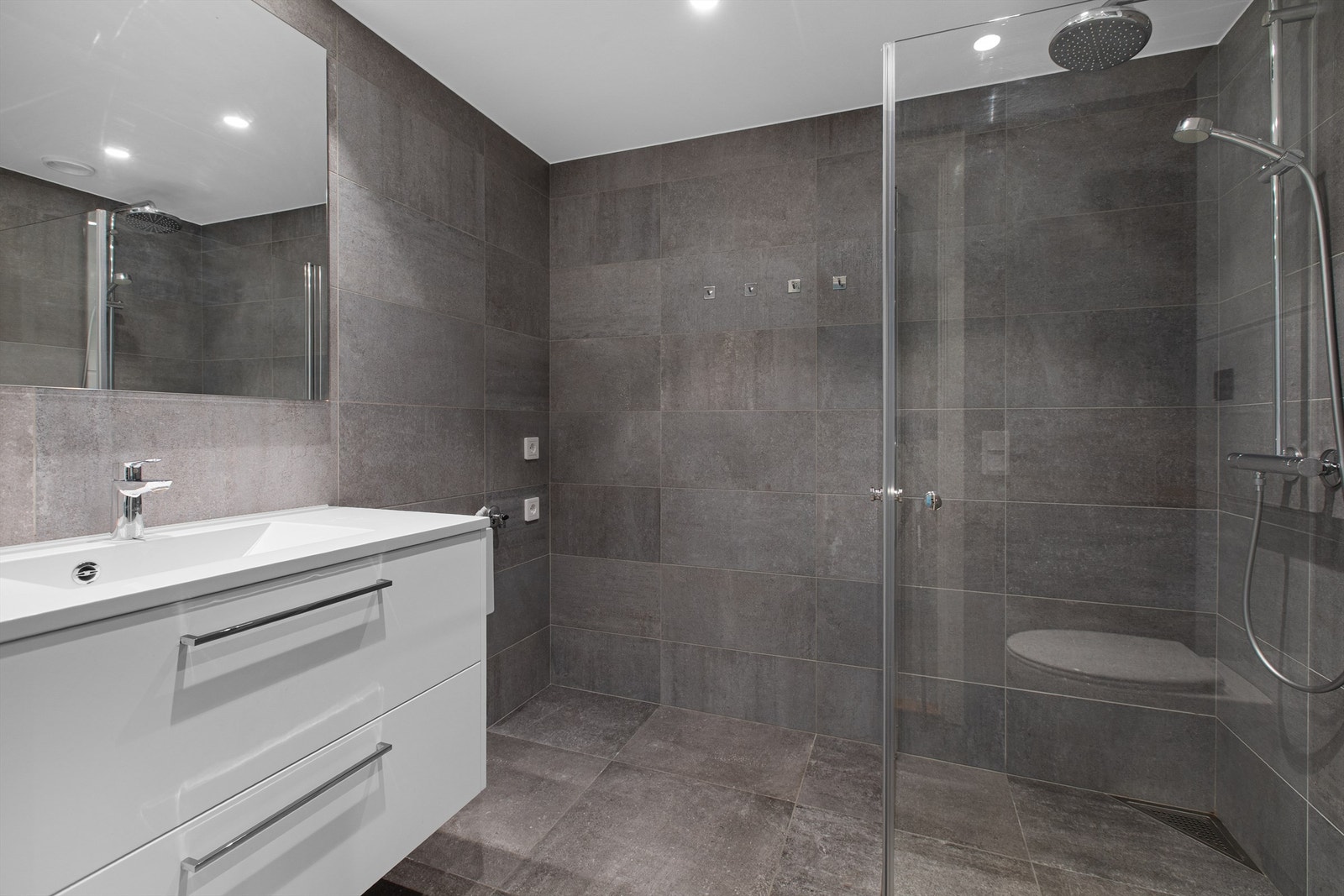 Badet har flislagt gulv med gulvvarme. Flislagte vegger. Malt himlingflate med downlights. Vegghengt servantskap med heldekkende servant og ett-greps armatur. Dusjnisje med innfellbare glassdører og dusjarmatur. Galleribilde