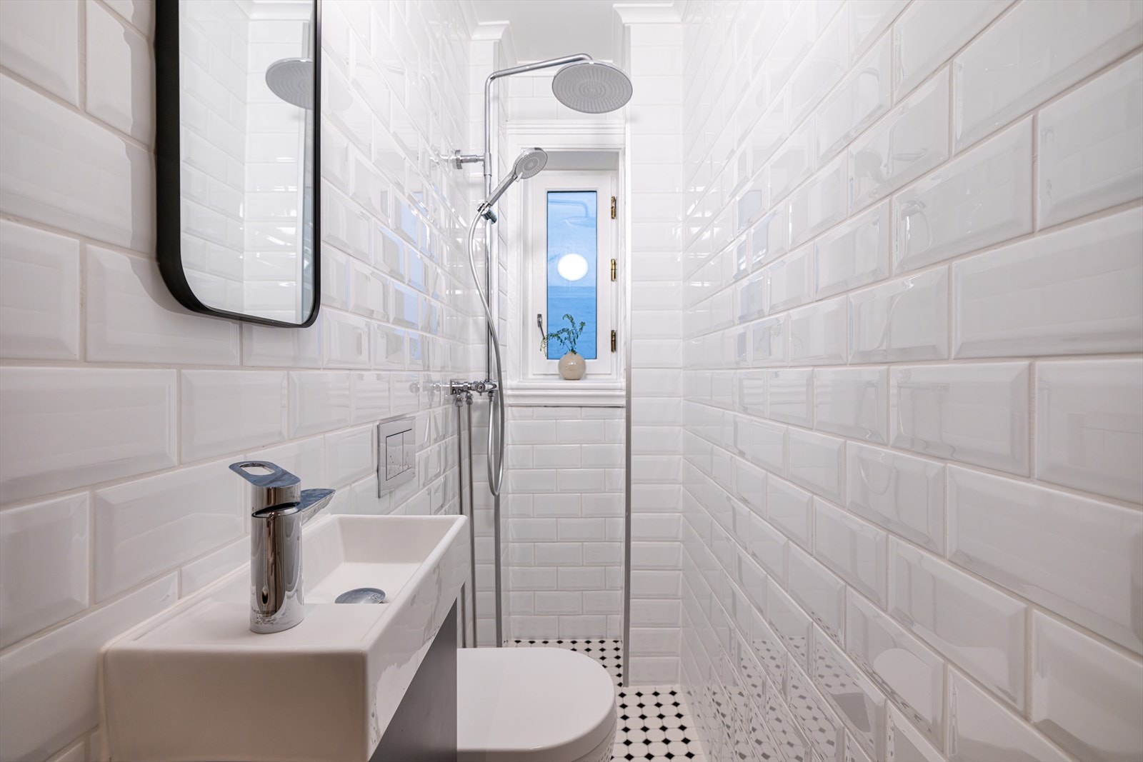 Flislagt bad med innredning bestående av benkeskap, speil over servant, dusj på gulv og veggfestet klosett med innebygget sisterne Galleribilde