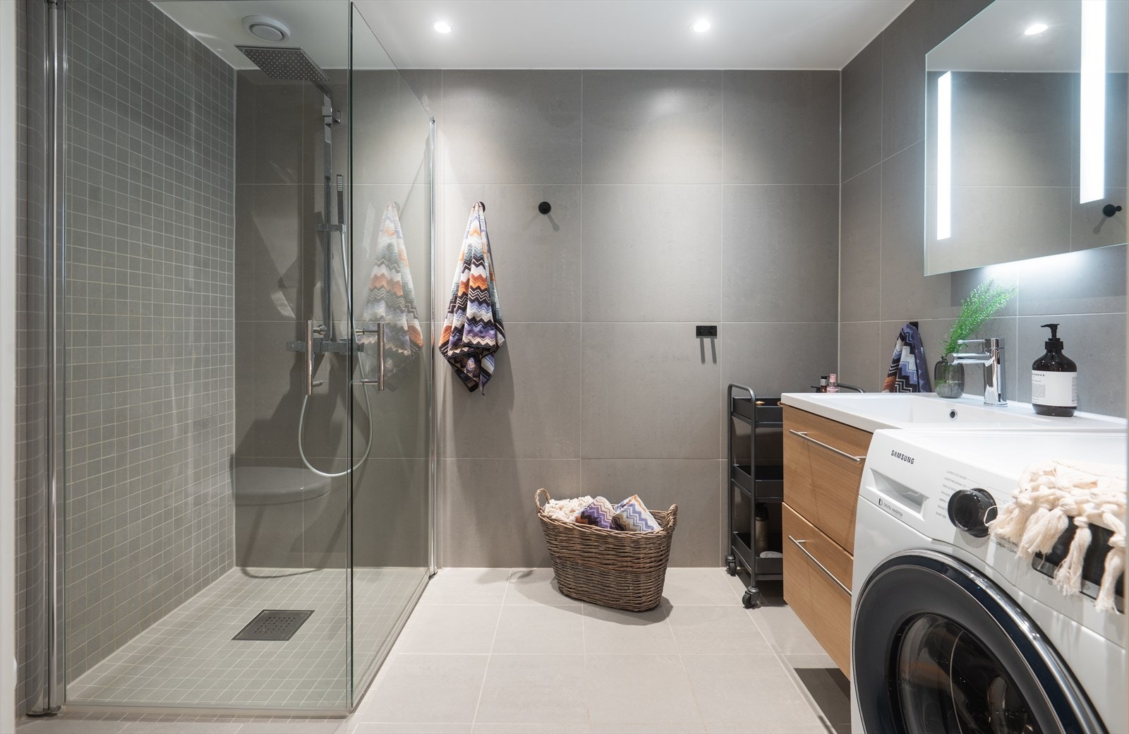 Bad med nedsenket dusjhjørne med dører i herdet glass, vegghengt toalett, servant med underskuffer og speil med LED lys over servant. Galleribilde