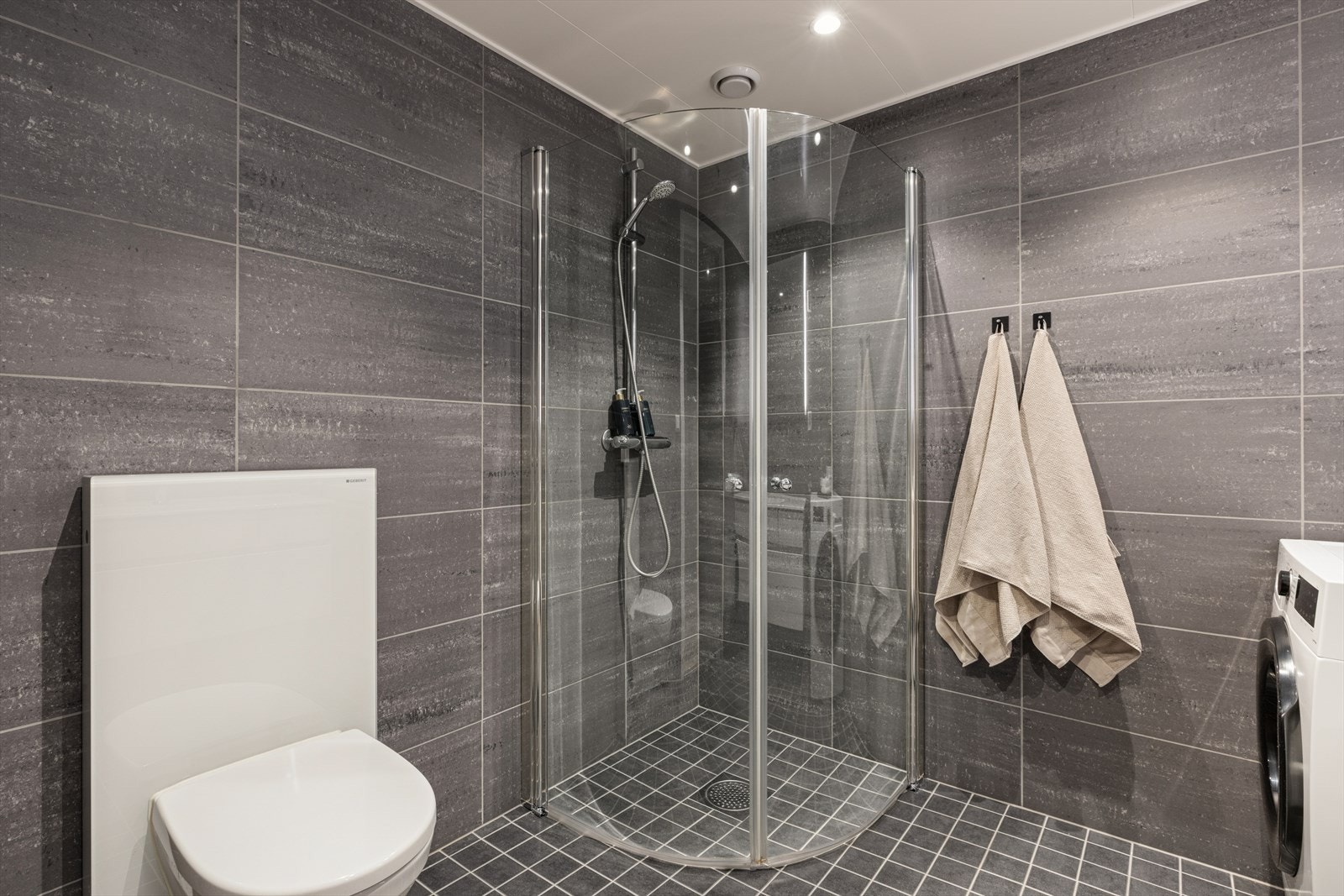 Badet er utstyrt med vegghengt toalett, dusjhjørne med svingbare glassdører, samt baderomsinnredning med nedfelt servant og speil. Galleribilde