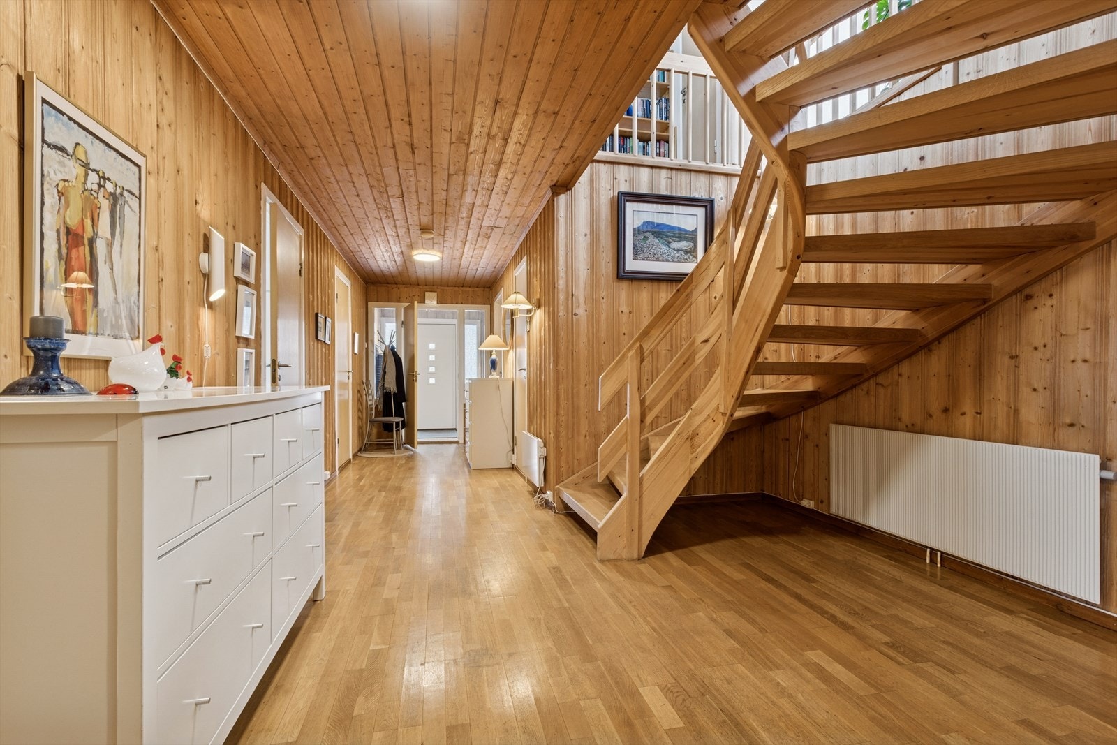 Videre inn i boligen kommer du til en romslig hall som fungerer som et naturlig knutepunkt i etasjen. Her finner du god plass til møblering etter behov, og fra hallen leder et åpent og oversiktlig trapperom deg videre opp til boligens hovedetasje. Galleribilde