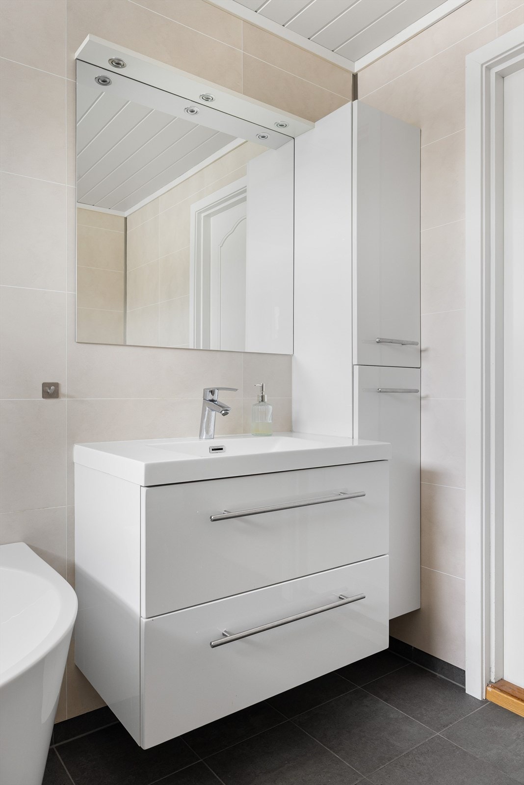 Baderomsrinnredning med skuffer og heldekkende servant. Over servanten er det speil med lys. Veggskap ved siden av. Galleribilde