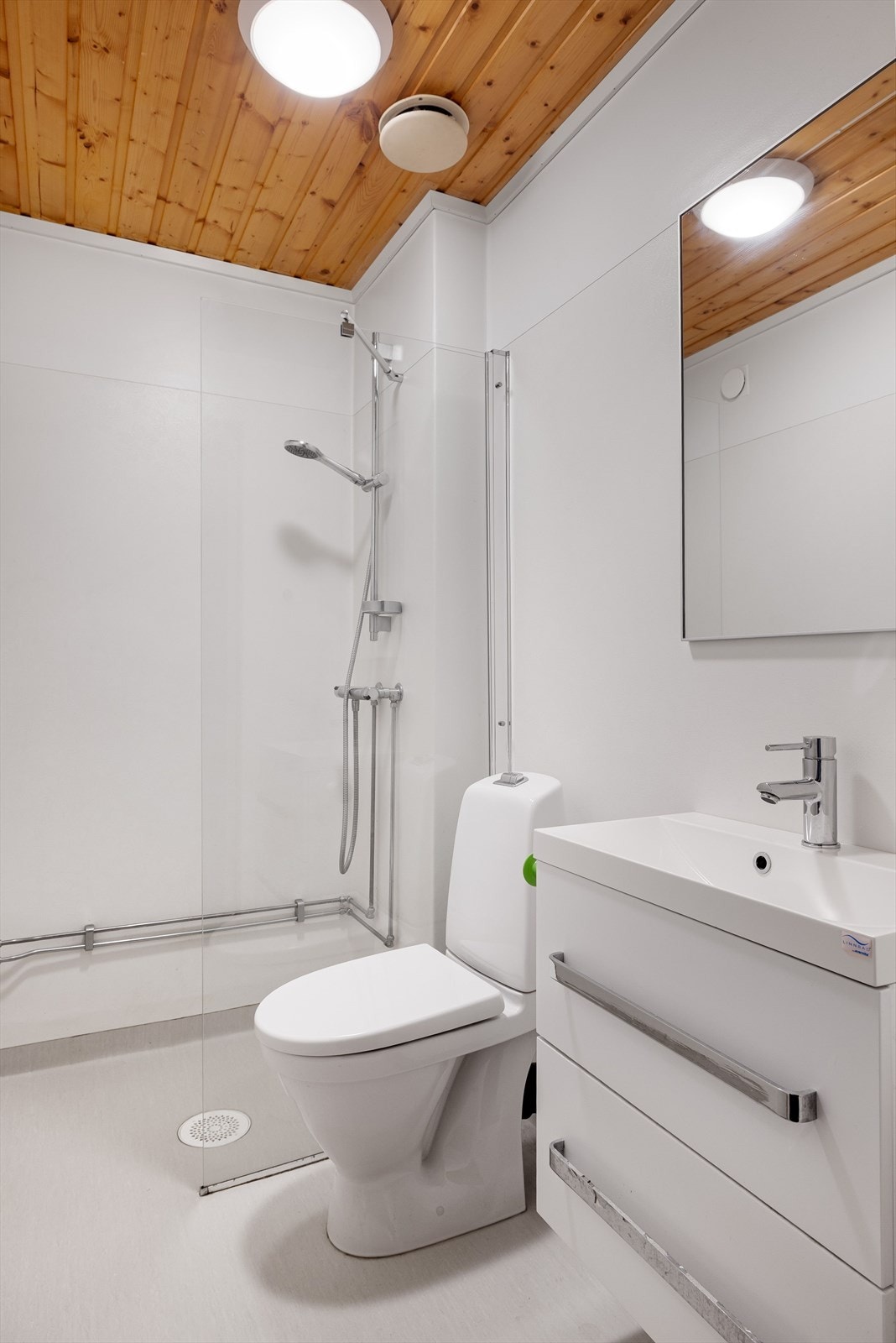 Bad i kjeller med vinylbelegg på gulvet og våtromstapet på veggene. Badet er utstyrt med gulvmontert toalett, baderomsinnredning og dusjnisje med veggmontert dusjbatteri og skillevegg. Baderomsinnredningen har fått nye knotter etter at bildet er tatt. Galleribilde