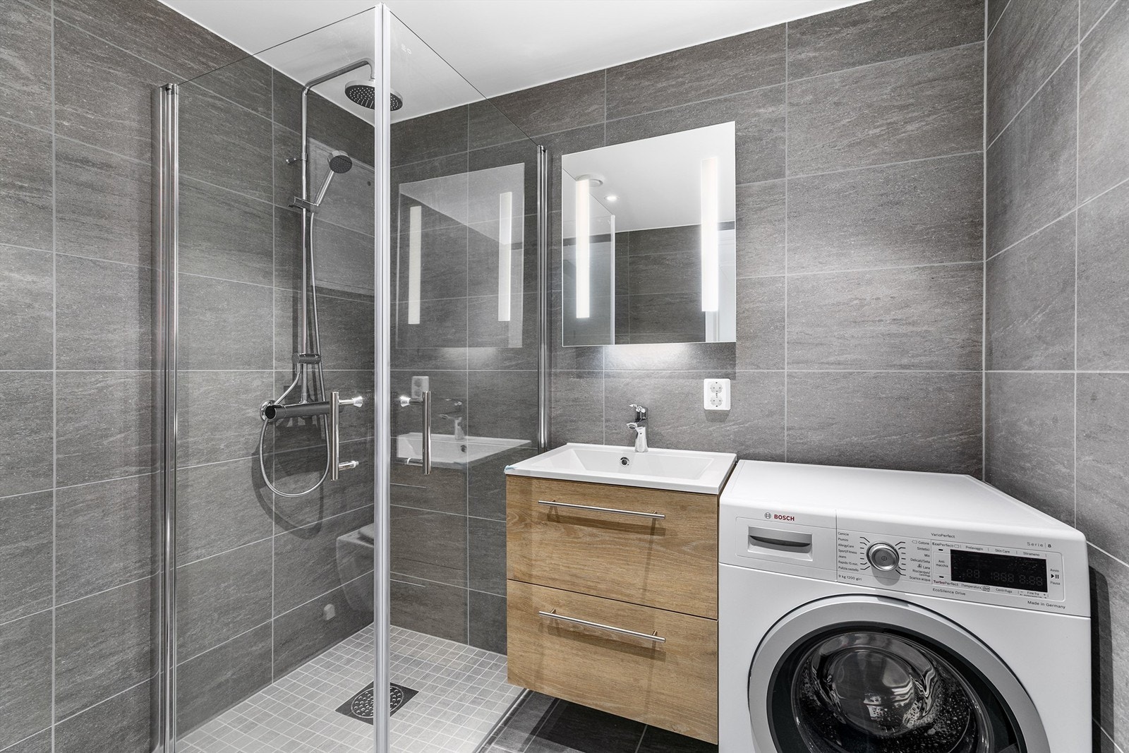 På badet er gulv og vegger utført med fliser i grå nyanser, i gulvet er det naturligvis også vannbåren varme. Legg merke til at vaskemaskinen medfølger. Galleribilde