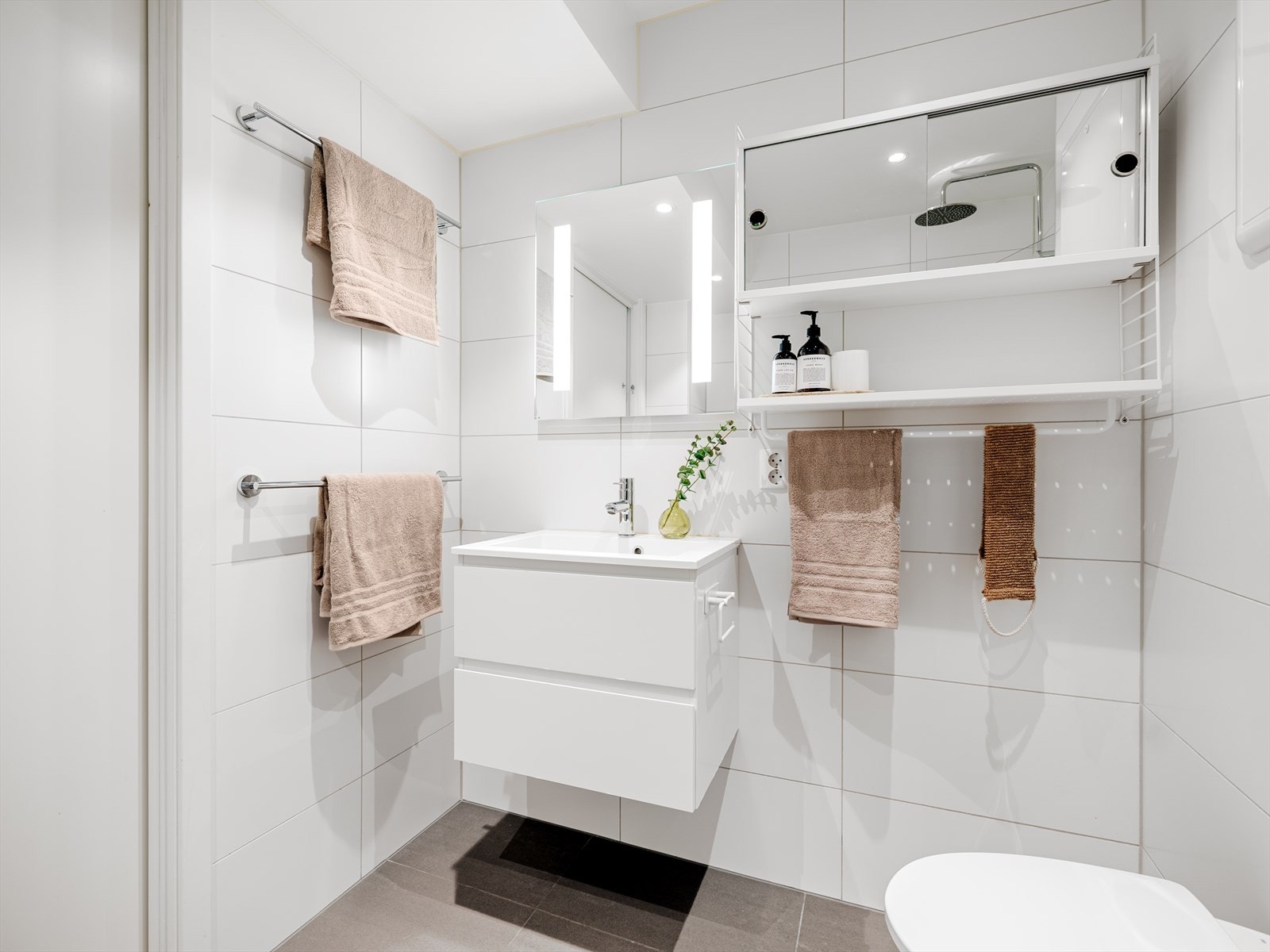 Badet er innredet med vegghengt toalett, baderomsinnredning med lysspeil, veggskap og dusjhjørne med regnfallsdusj Galleribilde