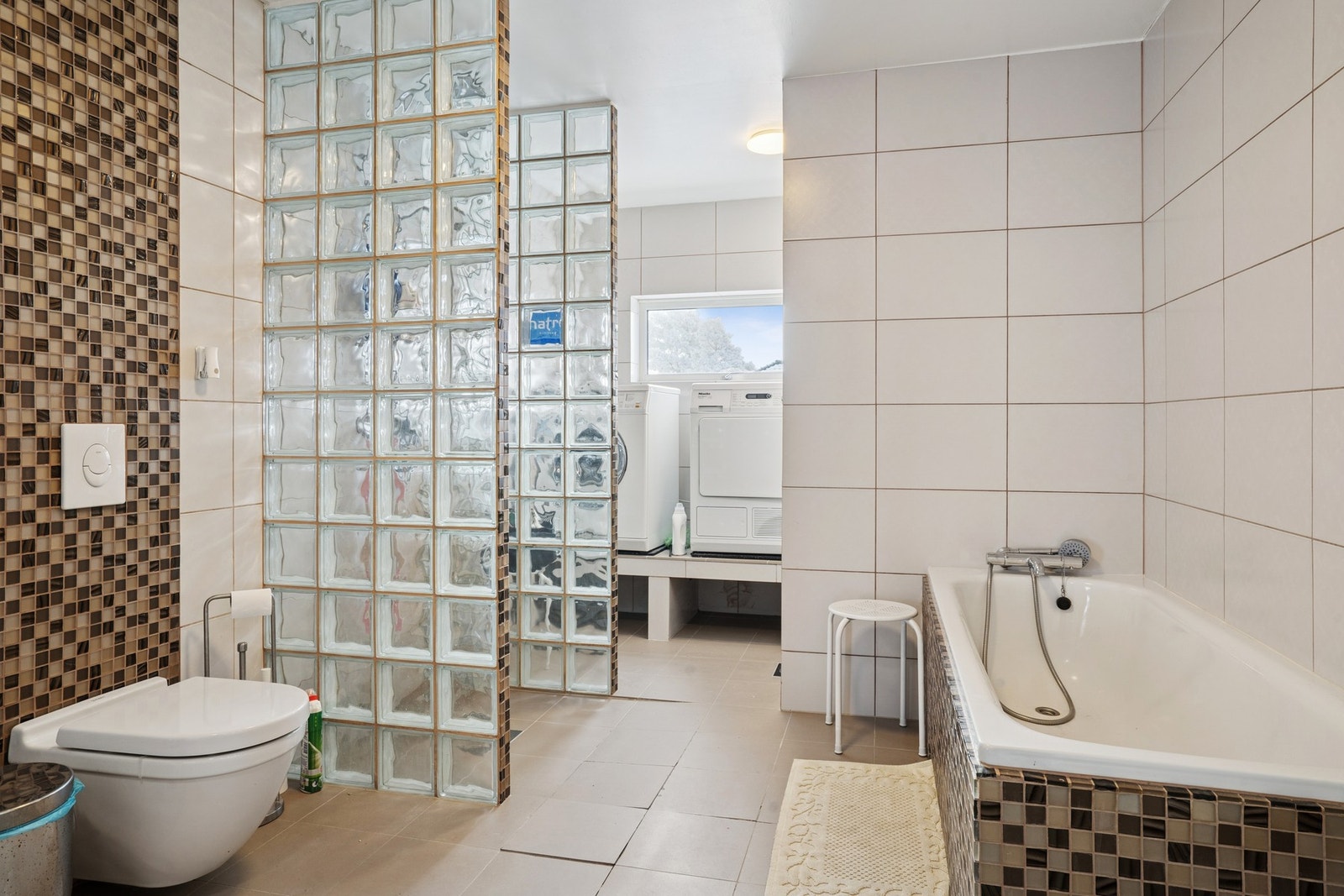 Innredet med servant med underskap, speilskap, gulvstående toalett, badekar og dusjløsning med glassbyggerstein. Galleribilde