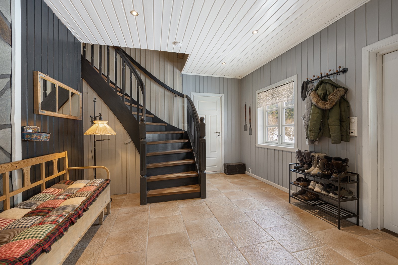 Her er det gråmalt panel på veggene, downlights i himlingen og beige fliser på gulv med gulvvarme. Det er god plass til garderobeløsning, og under trappen til 2. etasje er det dør til kjelleren. Galleribilde