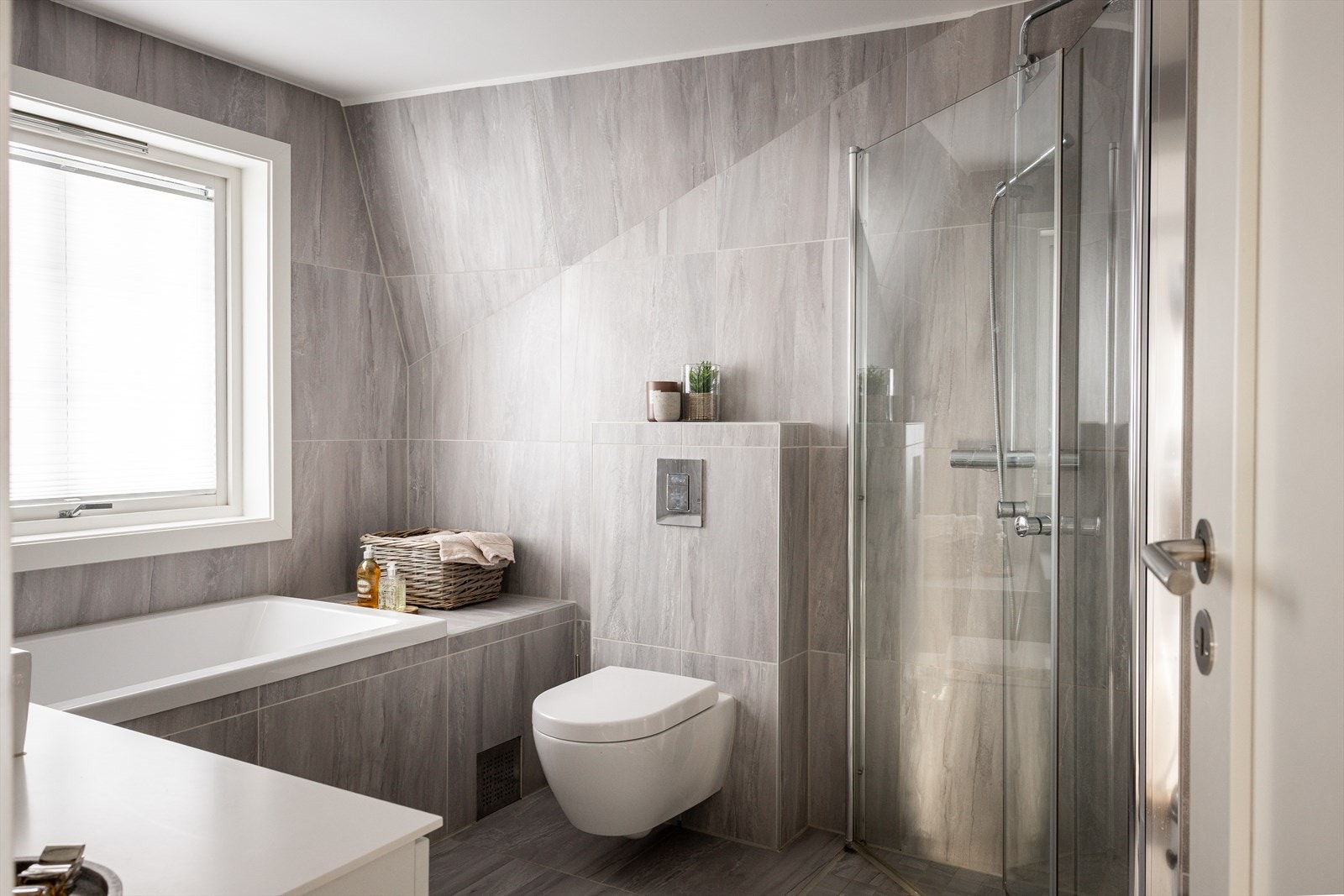 Bad med dusj med innfellbare dører i herdet glass, servant, veggfestet klosett med innebygget sisterne, og innfliset badekar. Galleribilde