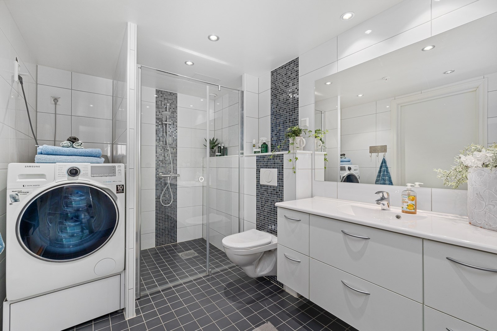 Baderommet er funksjonelt og stilrent innredet med dusjhjørne, baderomsmøbel med servant og speil, vegghengt toalett, samt en praktisk nisje med opplegg for vaskemaskin. Galleribilde