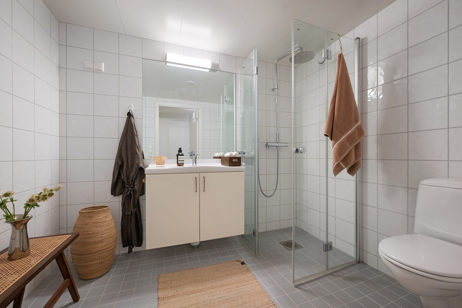Pent flislagt bad med gulvvarme og opplegg for vaskemaskin. Badet er utstyrt med toalett, dusj med innfellbare dører i herdet glass, benkeskap med glatte fronter og servant i helstøpt plate. Galleribilde