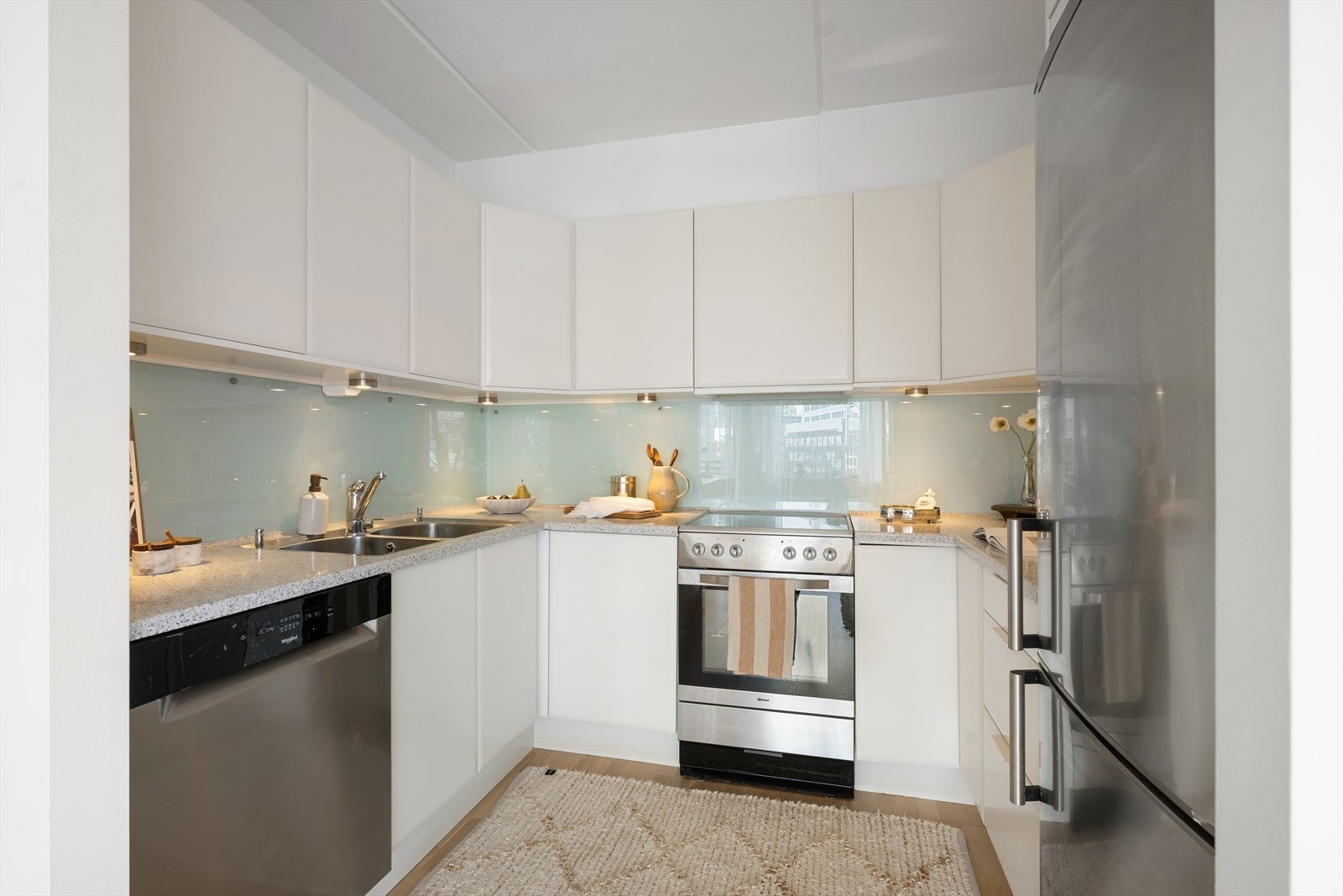 Lys og stilren kjøkkeninnredning bestående av glatte fronter og laminat benkeplate med nedfelt kum i rustfritt stål. Det er lagt glassplater på vegg over benkeskap. Galleribilde