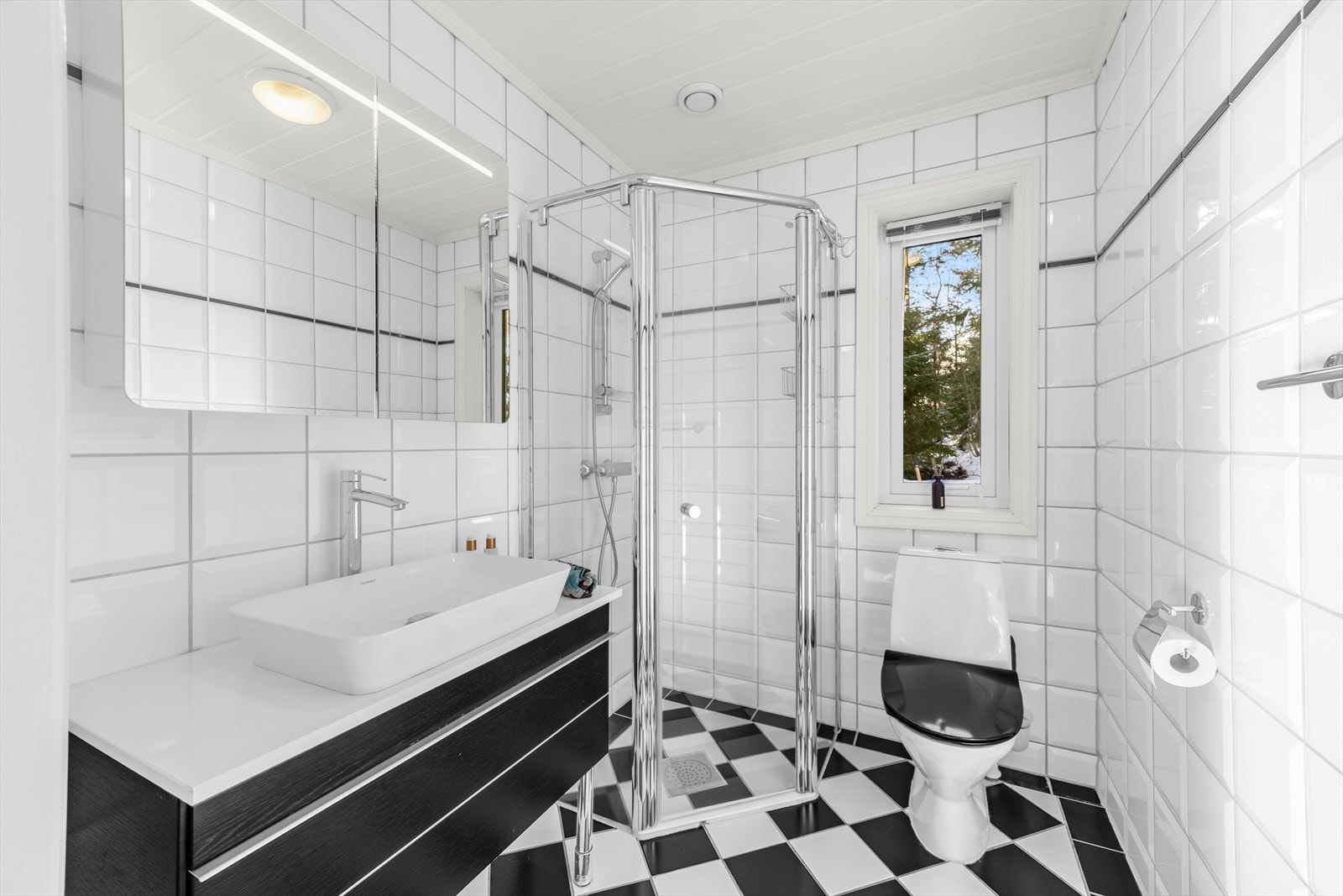 Badet i 1. etasje er pent med en kombinasjon av sorte og hvite fliser med gulvvarme. Innredet med veggmonterte dusjvegger, dusj direkte på gulv med termostat batteri, klosett, servant med mørk skuffinnredning og speilskap med belysning. Galleribilde