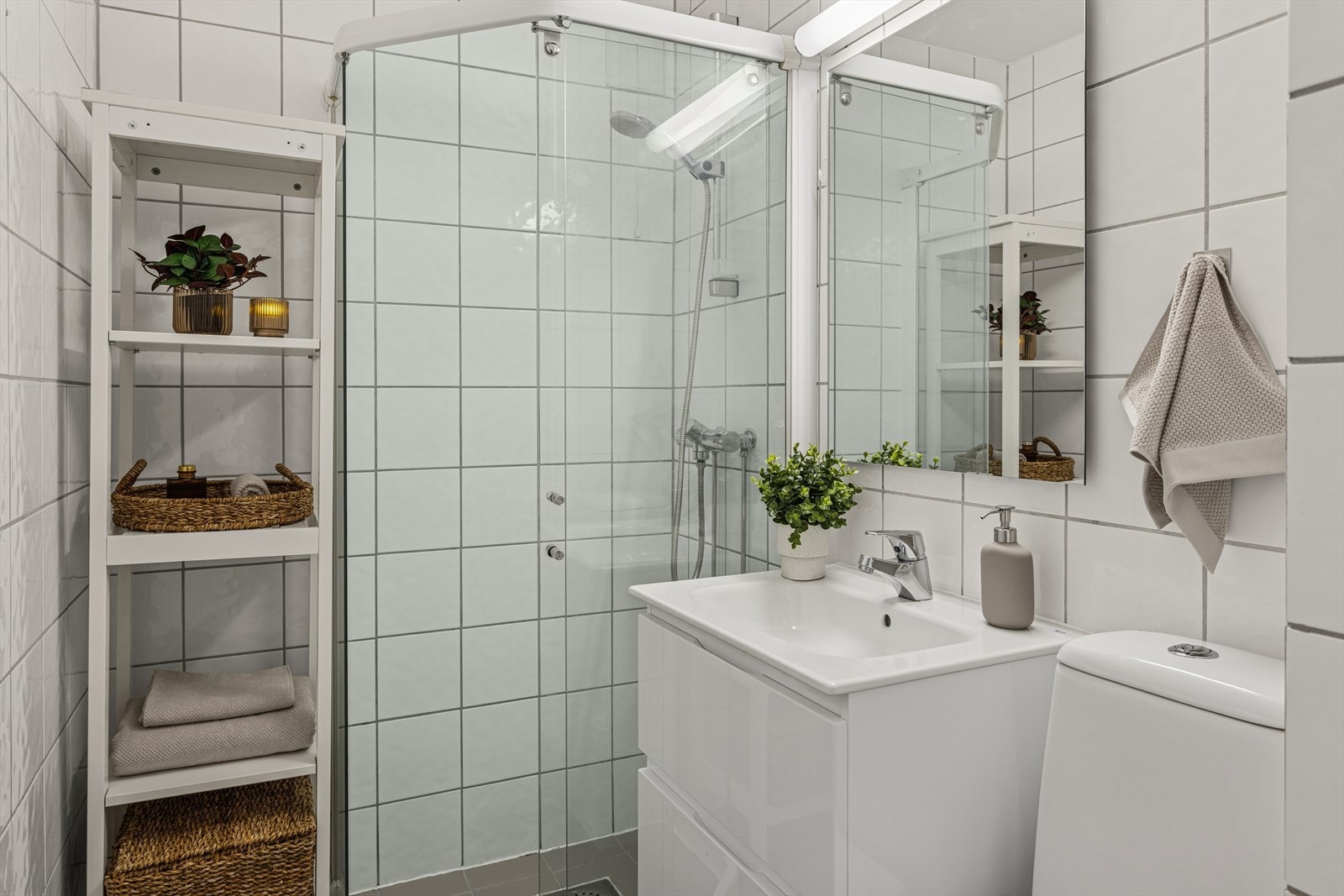 Et lyst baderom med fliser på gulv og vegger. Videre er baderommet innredet med klosett, servant med innredning, speil og dusjhjørne med skyvbar glassdør. Galleribilde