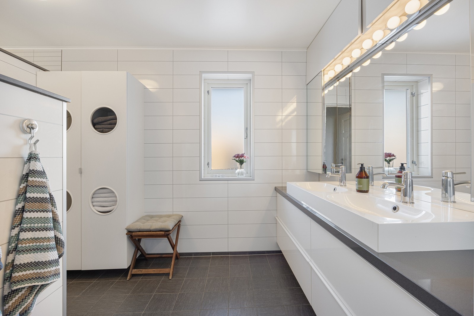 Badet fremstår romslig, praktisk og pent. Veggene har hvite våtromsplater, mens det på gulvet er grå fliser med lun varme under. Galleribilde