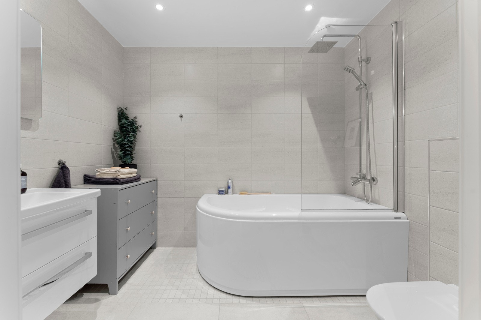 Stilfullt innredet med servantinnredning, speil med lys på vegg over servant, vegghengt toalett og badekar med dusjvegg i glass. Galleribilde