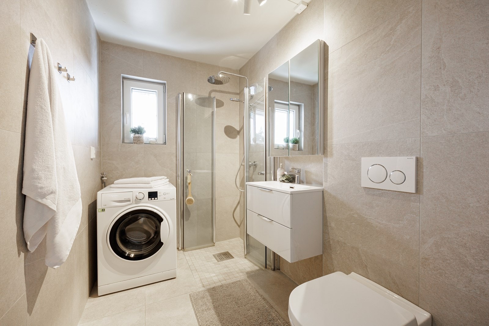 Flislagt bad med moderne innredning bestående av benkeskap med glatte fronter og veggskap med speilfronter. Vegghengt toalett med innebygget sisterne, dusj med innfellbare glassdører samt opplegg for vaskemaskin. Galleribilde