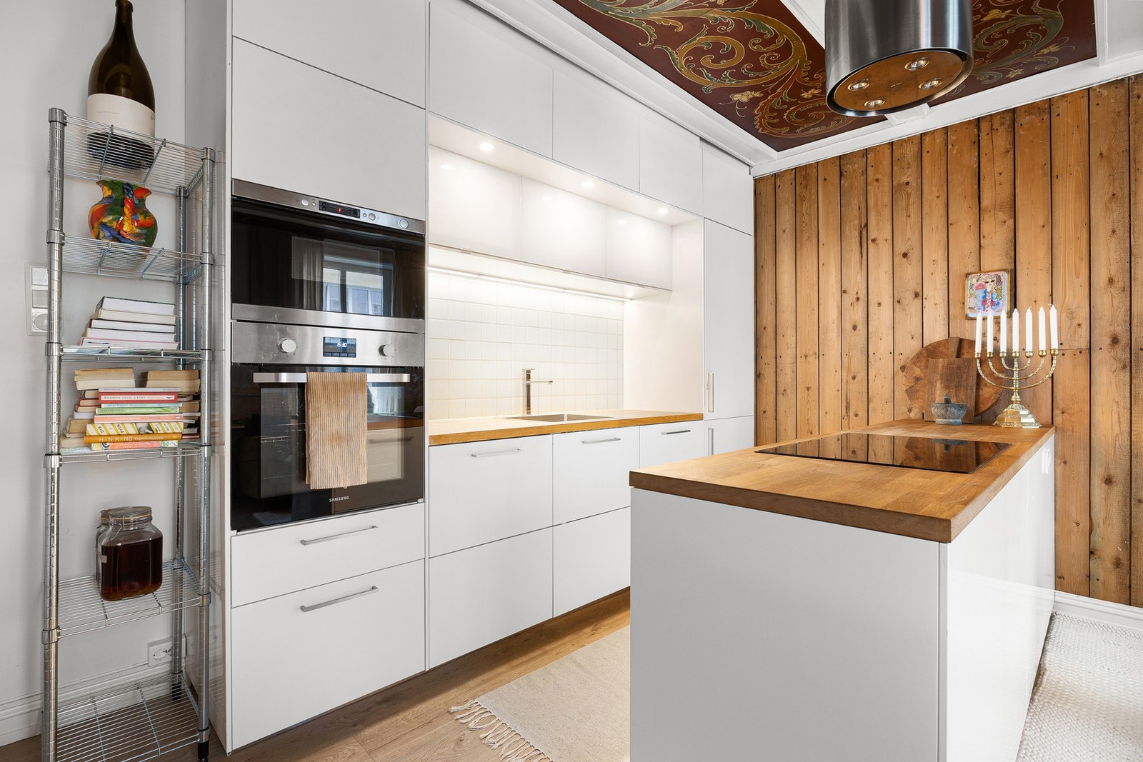 Fargepaletten er gjennomført til fingerspissene med en lun fargetone på vegg kombinert med detaljer av panel, takbjelker og rosemalte plater antatt fra byggeår Galleribilde