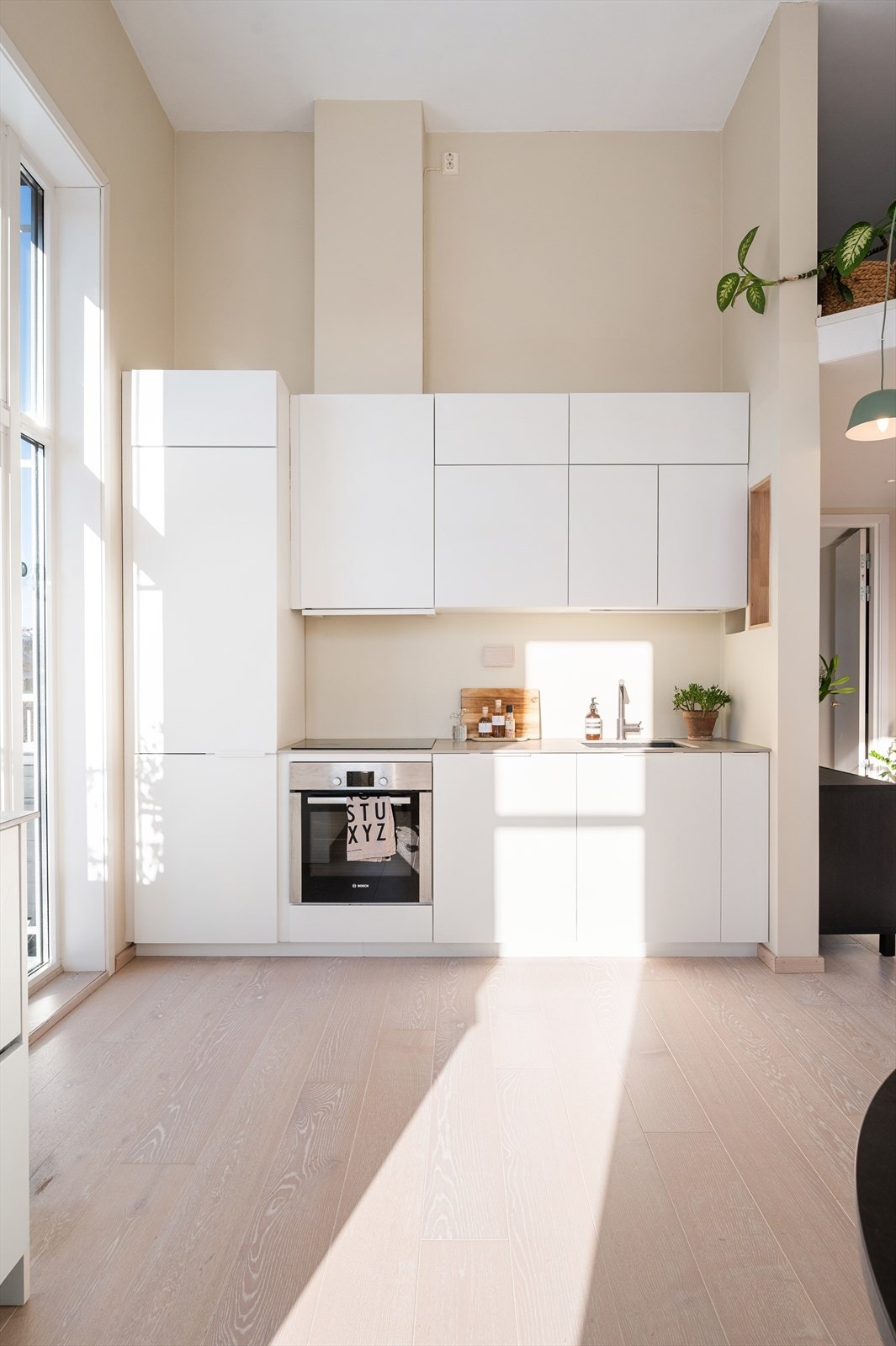 Kjøkkeninnredning fra KVIK med glatte fronter og belysning under overskap. Integrerte hvitevarer inkluderer induksjonstopp, stekeovn, oppvaskmaskin og kjøl/frys. Galleribilde