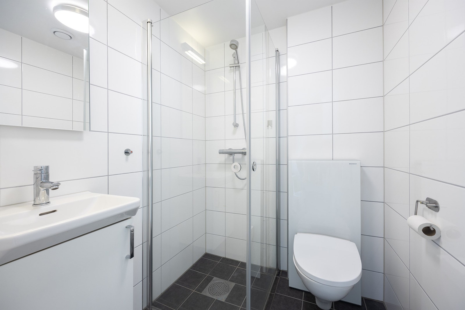 Badet fra soverom III / hybeldelen er lyst og hyggelig med veggmontert toalett og speil, heldekkende servant med underskap og dusjhjørne med dører i herdet glass Galleribilde