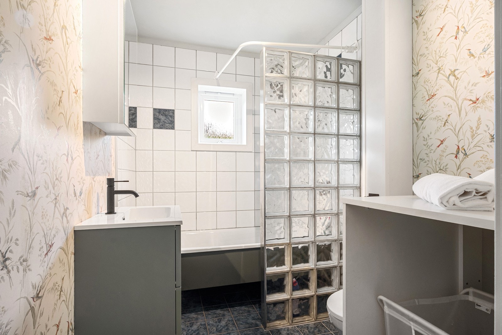 Badet har flislagte overflater i dusjsonen, sokkelfliser med varmekabler og en dekorativ tapet på vegger. Innredning består av servant med skuffer, speil, toalett og badekar med dusjmulighet. Galleribilde