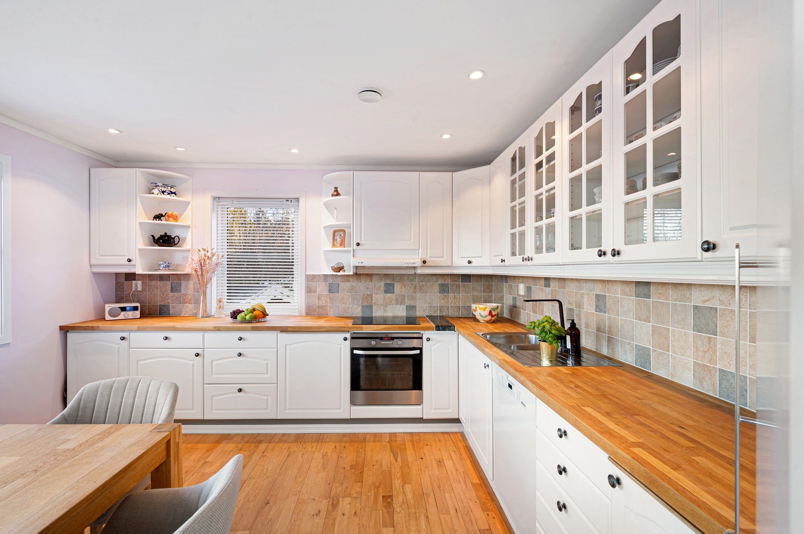 Kjøkkeninnredning med over- og underskap, profilerte møbelplater i fronter og heltre benkeplater. Ventilator over stekesone. Integrerte hvitevarer som platetopp og stekeovn. Kran og avløp for oppvaskmaskin. Galleribilde