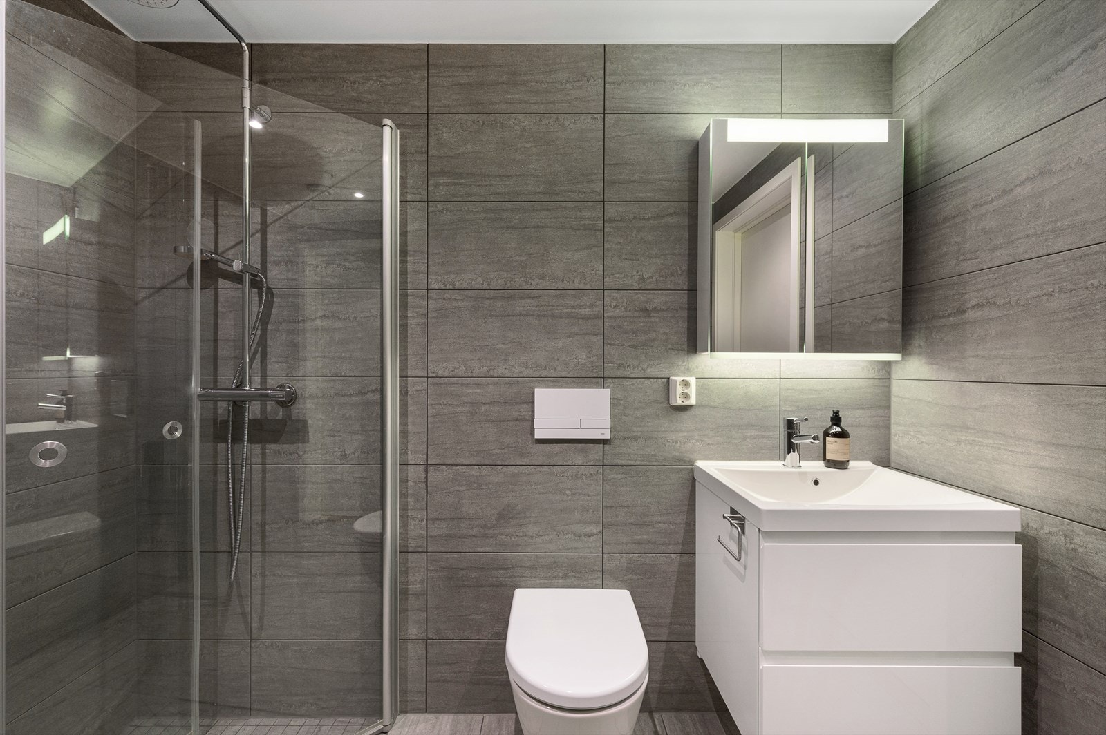 Badet er innredet med vegghengt toalett, dusjhjørne m/glassdører, opplegg for vaskemaskin, samt servant med innredning, speilskap og belysning. Galleribilde