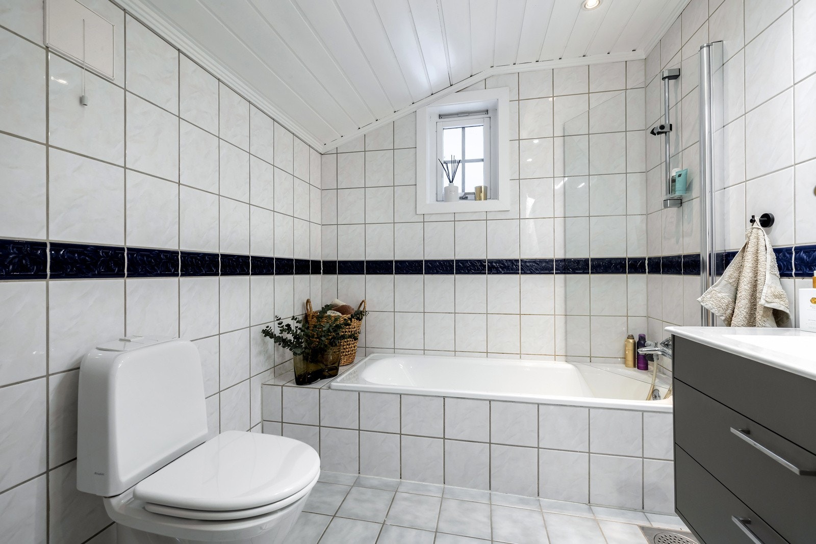 Badet er innredet med servant i moderne baderomsinnredning med skuffer, speil med belysning, toalett samt badekar med dusjløsning og glassvegg. Det er også høyskap som gir ekstra oppbevaringsplass. Galleribilde