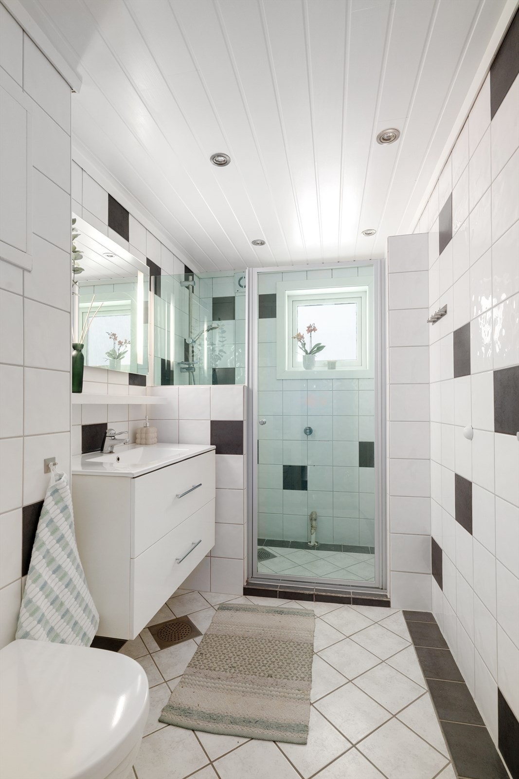 Flislagt baderom fra 2006 som inneholder heldekkende servant med underskap, speil, veggmontert toalett og dusjnisje med glassdør. Galleribilde