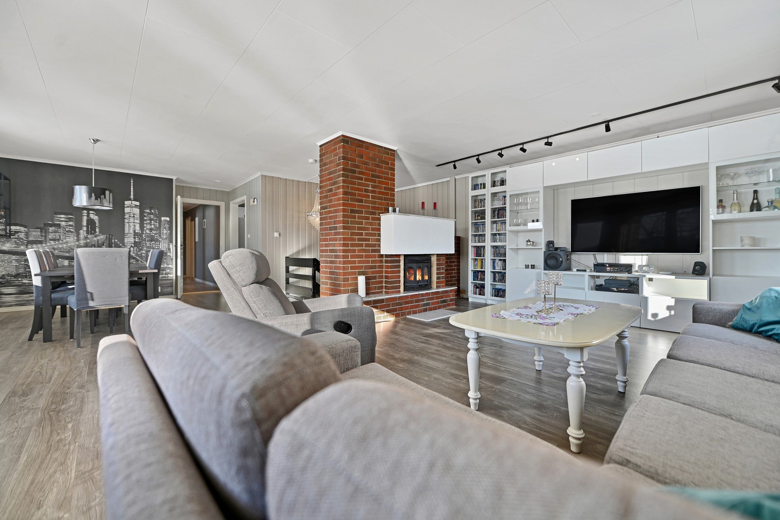 Stue med peis, åpen løsning mot spiseplass, og innebygde hyller med TV. Lysmalte vegger og parkettgulv. Galleribilde