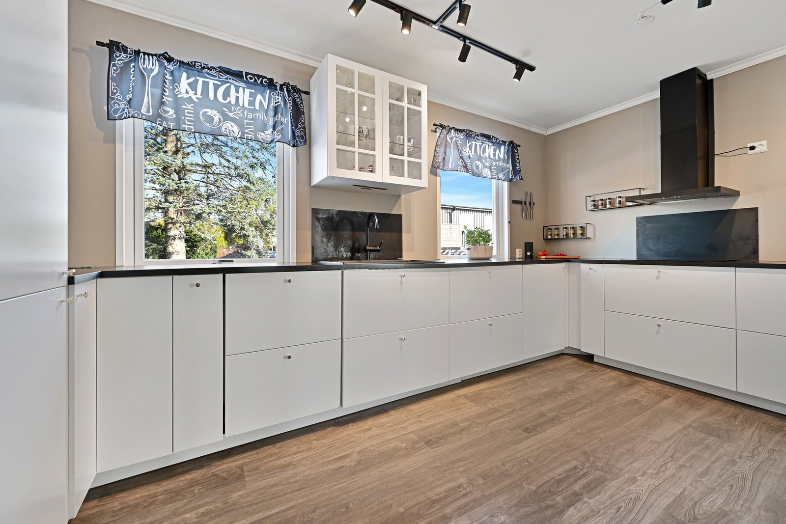 Lyst kjøkken med moderne hvite skap, mørk benkeplate og store vinduer som gir godt med naturlig lys. Galleribilde