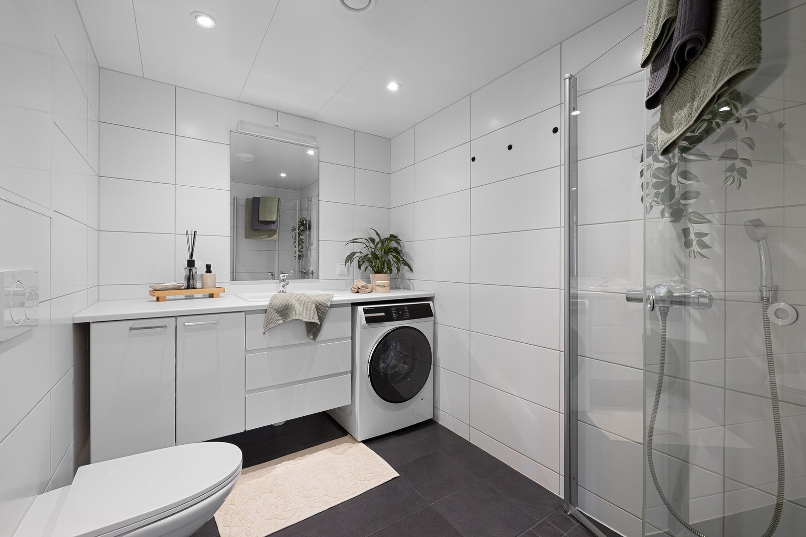 Leilighetens bad, med fliser på både gulv (elektriske varmekabler) og vegger, fremstår lyst og innbydende. Utstyrt med innfellbare og plassbesparende dusjdører i glass, kran og avløp for vaskemaskin og vegghengt WC. Galleribilde