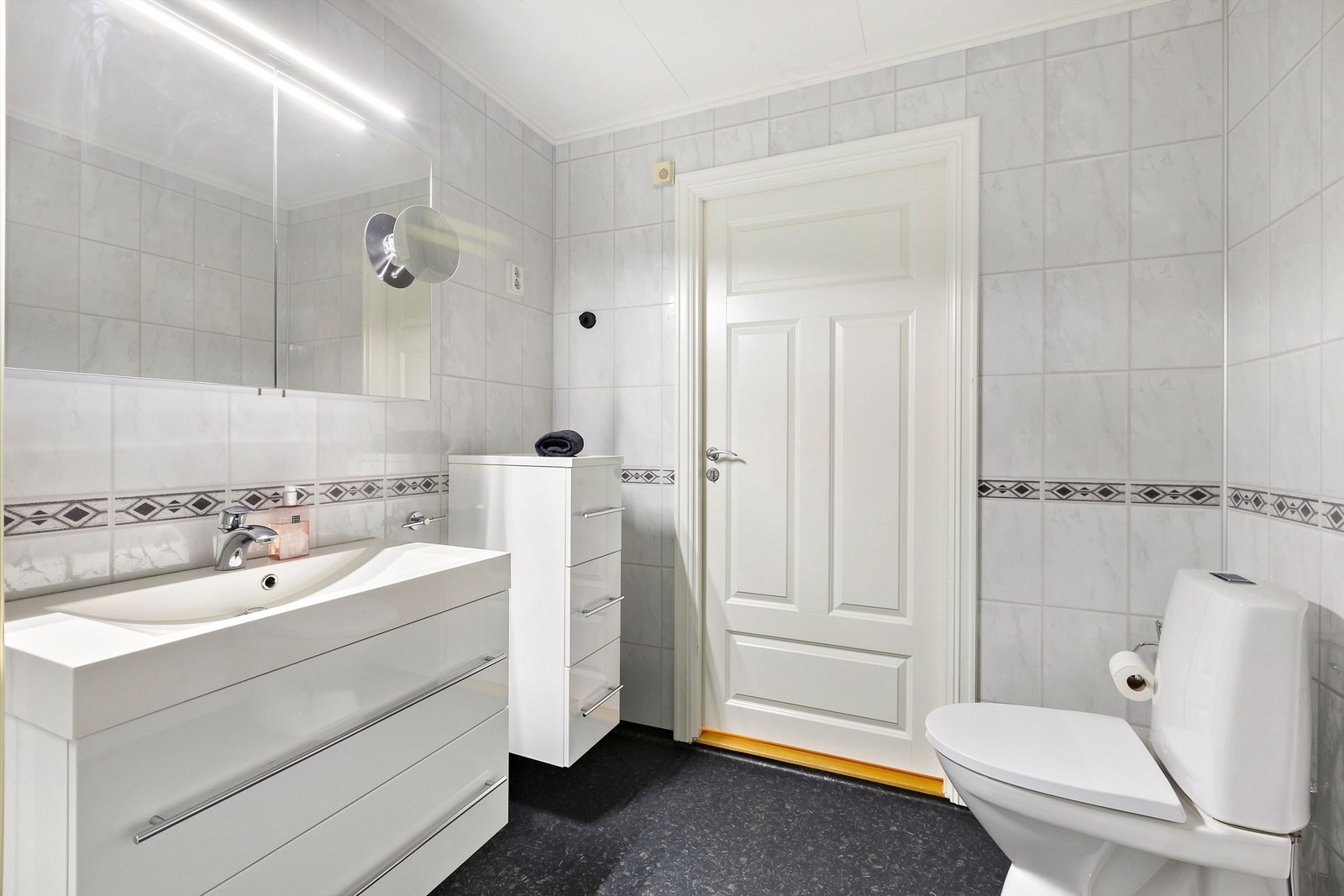 Badet i 1. etasje har hvit innredning med nedfelt servant, speilskap og gulvmontert toalett Galleribilde