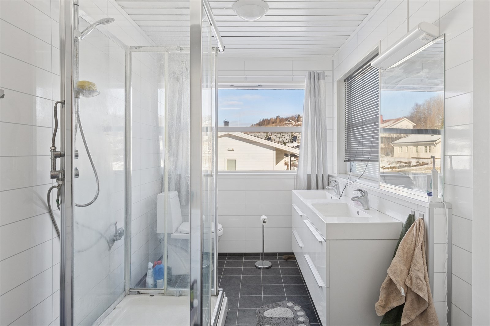 Flislagt bad. Badet er utstyrt med blant annet veggmontert dobbeltservant, frittstående dusjkabinett og opplegg for vaskemaskin. Galleribilde