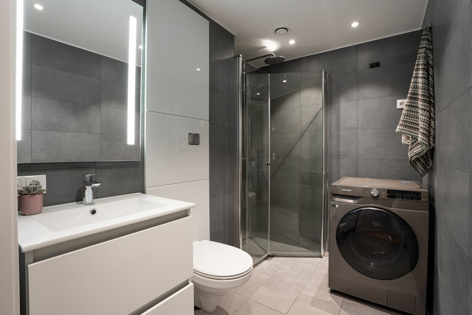 Leiligheten har et lekkert bad med moderne flisvalg og baderomsinnredning. Her er det vegghengt toalett, dusjhjørne og opplegg for vaskemaskin. Galleribilde