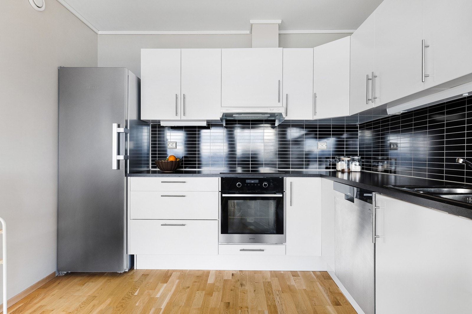 Kjøkkeninnredning i hvit utførelse med slette fronter, laminert benkeplate og mørk kontrastvegg over kjøkkenbenk. Integrert platetopp, stekeovn og oppvaskmaskin. Galleribilde