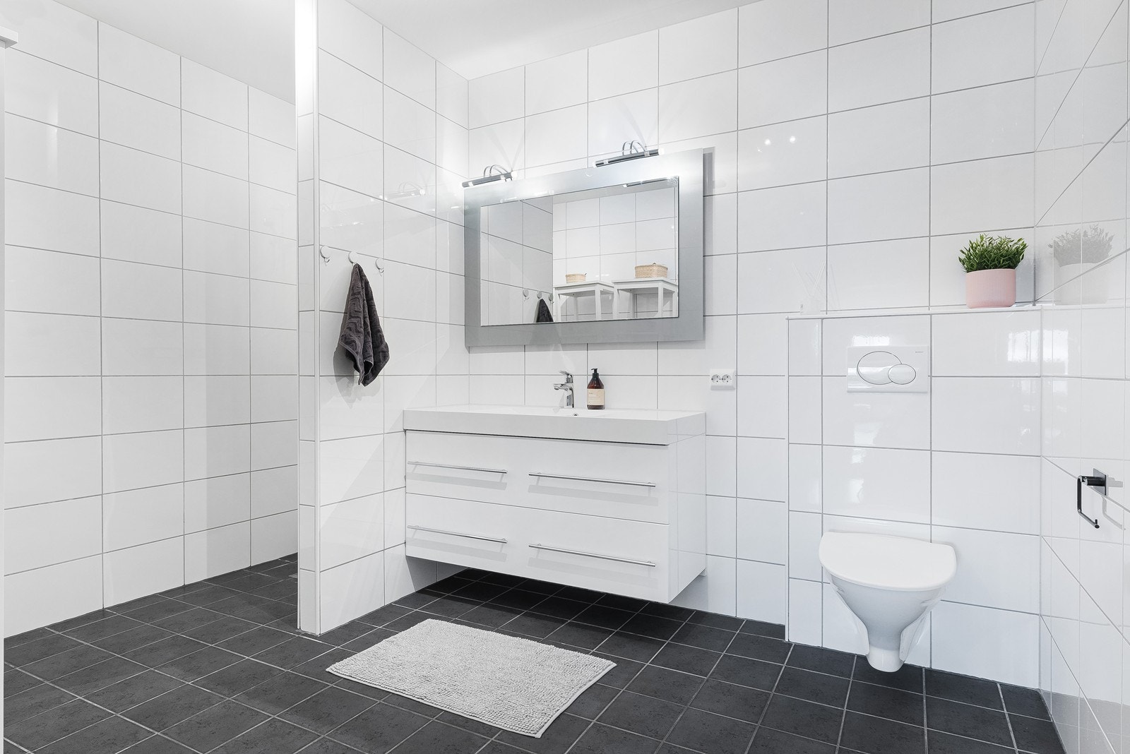 Baderom med fliselagt gulv og vegg og varmekabler, vask i dobbel skuffeseksjon med speil, vegghengt toalett og dusjhjørne. Galleribilde