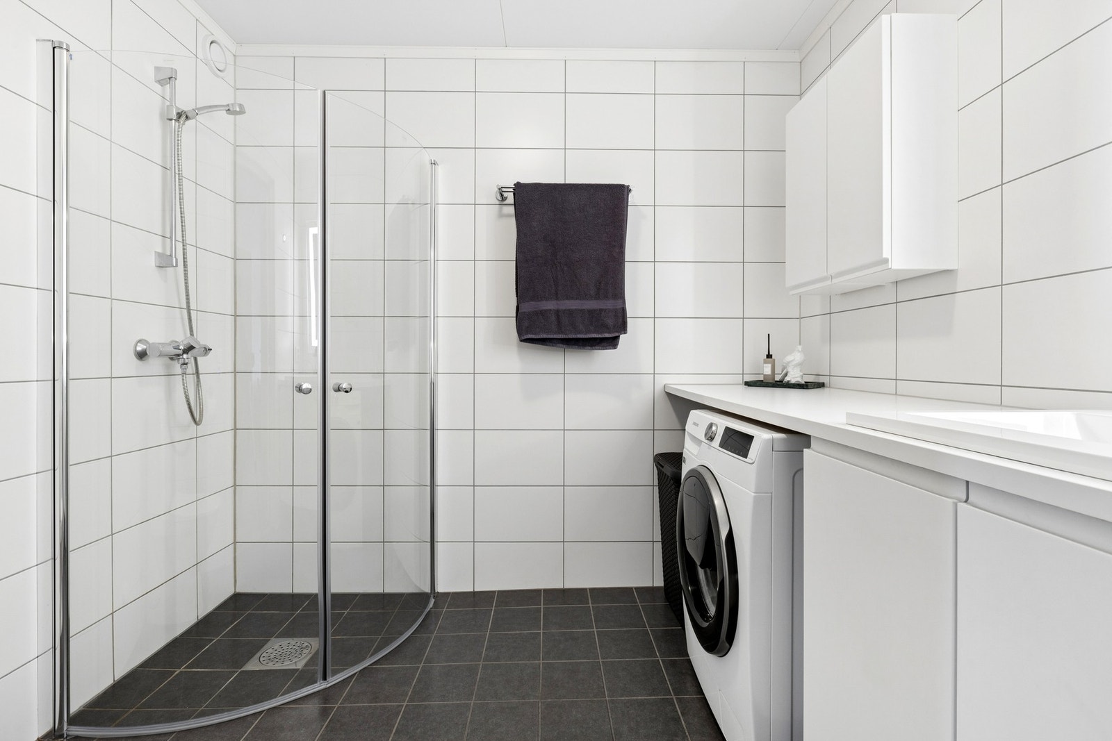 Badet er helfliset og praktisk utformet med dedikert plass til vaskemaskin, dusjhjørne med glassdører, servantinnredning med skap og gulvstående toalett. Galleribilde