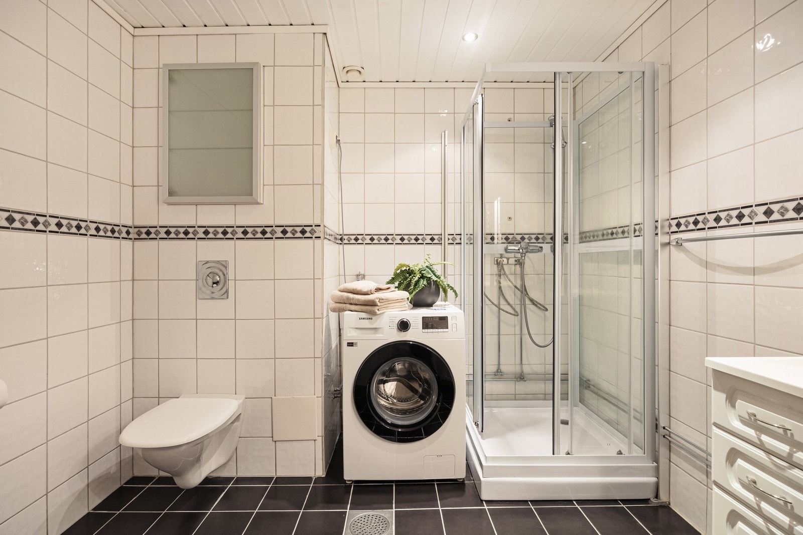 Badet er innredet med vegghengt servantinnredning med ovenpåliggende servant og armatur, speil med overlys og stikkontakt, vegghengt skap, dusjkabinett og vegghengt toalett. Galleribilde