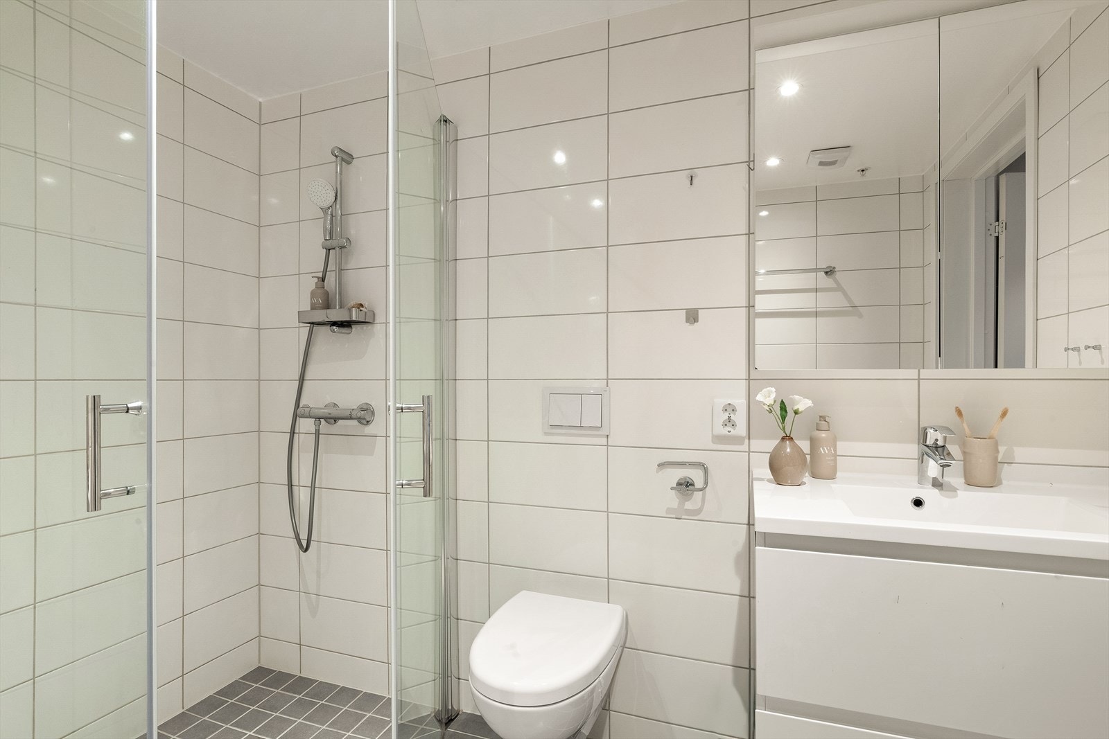 Baderomsinnredningen har fliser på vegg og gulv, servant med skuffer, speilskap, dusj med veggmonterte glassdører, vegghengt klosett og opplegg for vaskemaskin. Galleribilde