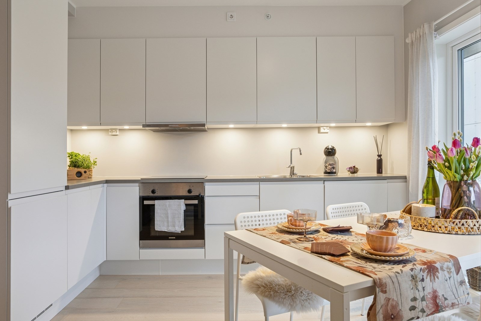 Spotter under overskap gir godt arbeidslys og setter en behagelig stemning i rommet. Store vinduer sørger for at både stuen og kjøkkenet fylles med naturlig lys, noe som skaper en åpen og innbydende atmosfære. Galleribilde